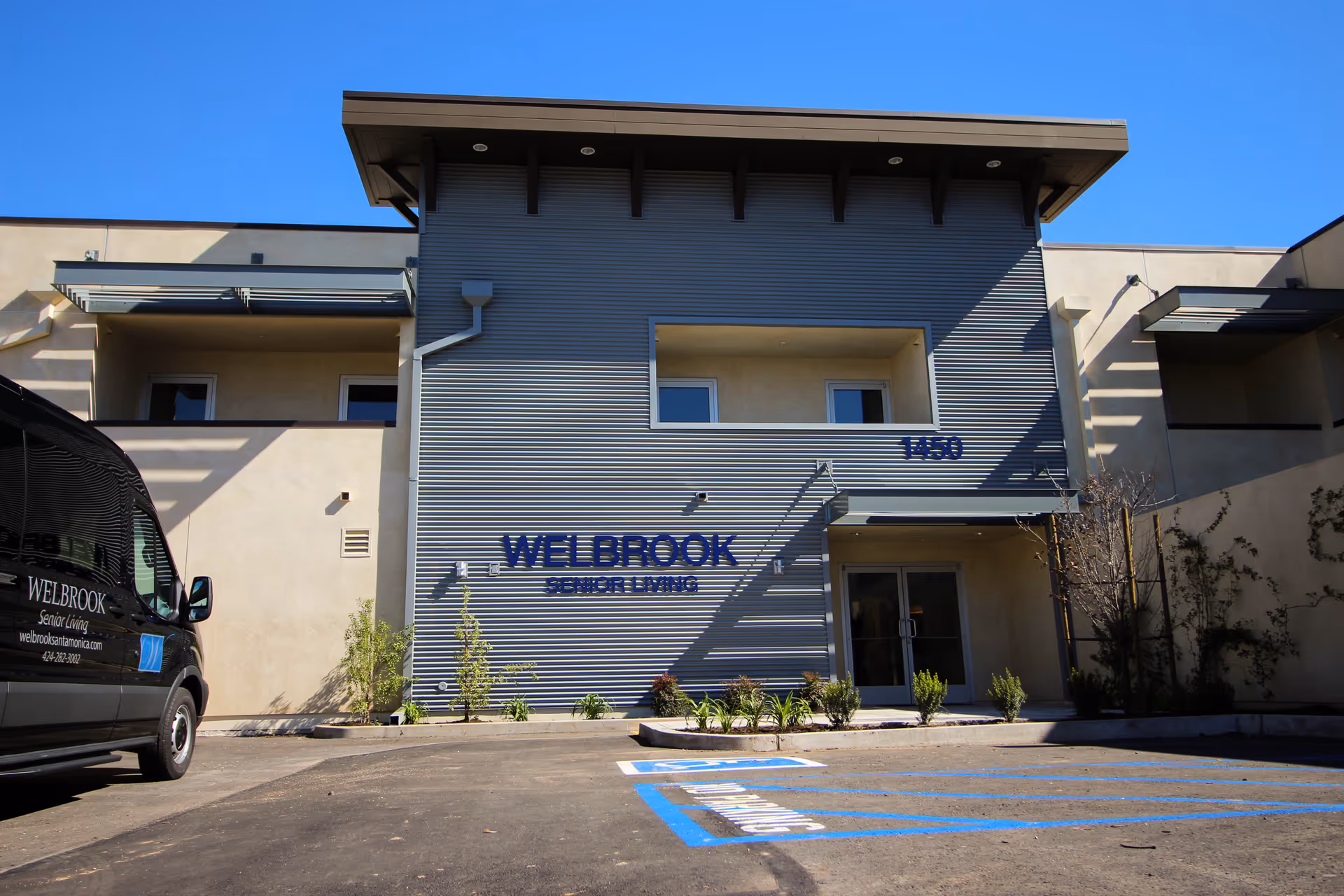 Front entrance of the Welbrook Senior Living building with its signage, parking spaces, and a facility van parked at the left.