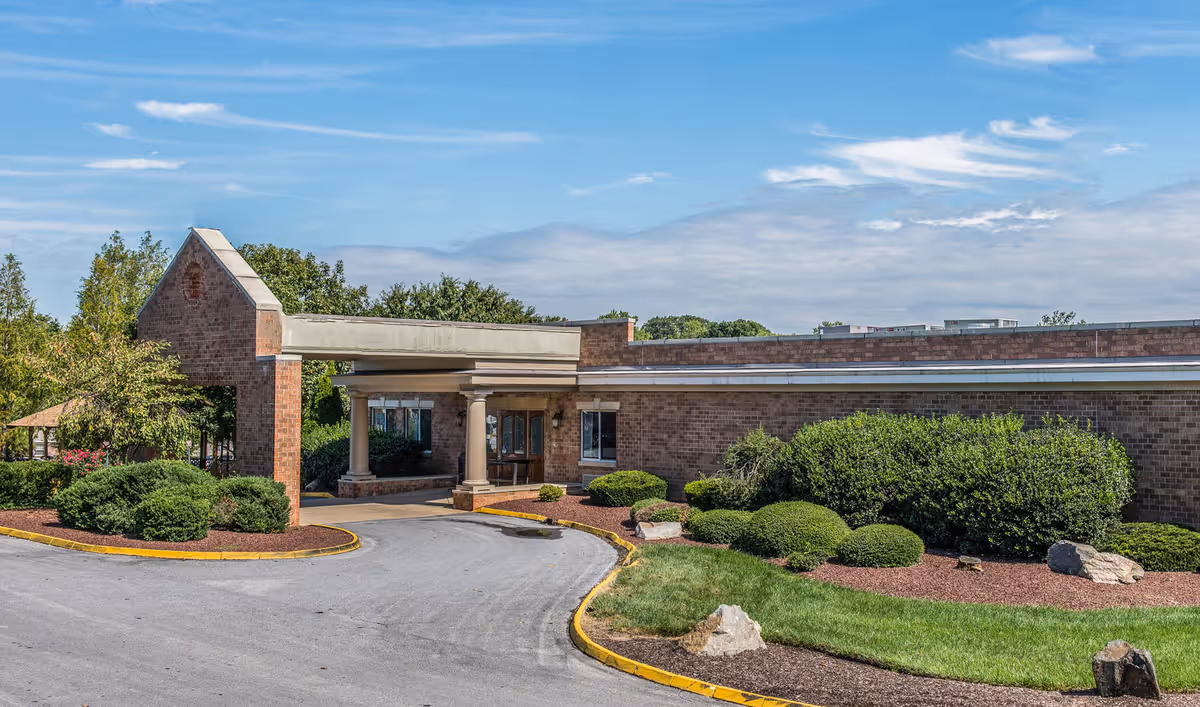 One-story brick senior facility with a covered drive-up entrance, driveway and landscaped shrubs under a blue sky.
