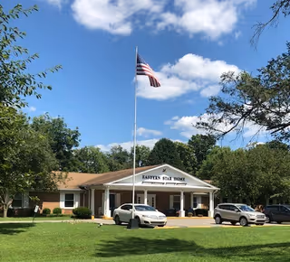 Front entrance of the Eastern Star Home building with a tall American flag, parked cars, and a grassy lawn under a blue sky.