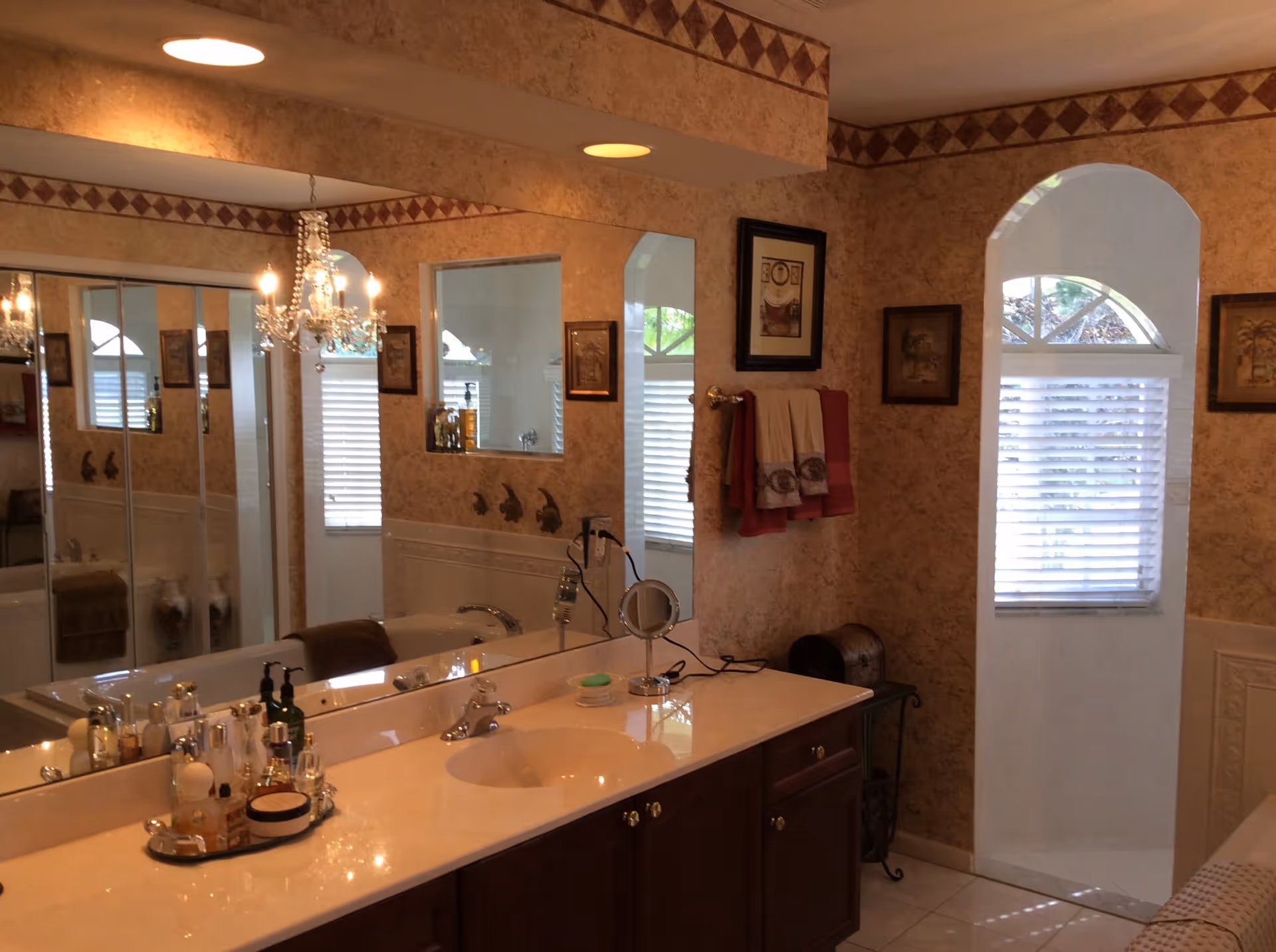 Bathroom with a long vanity and sink, large mirrored wall, chandelier, decorative wallpaper, and an arched window with blinds.