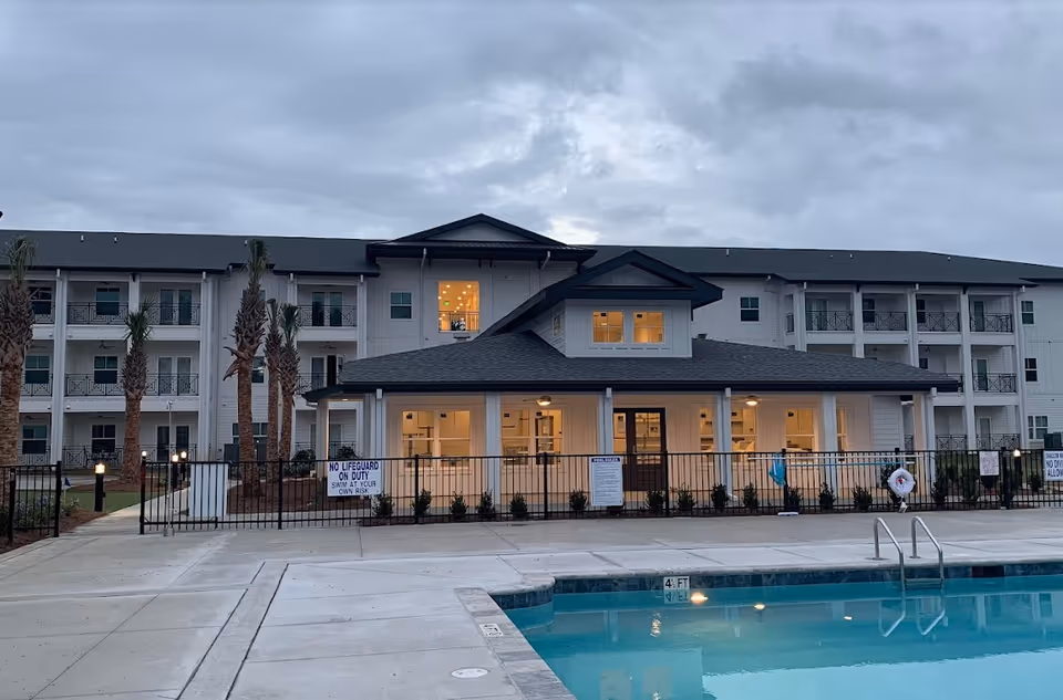 Three-story senior living building with a fenced pool in the foreground at dusk.