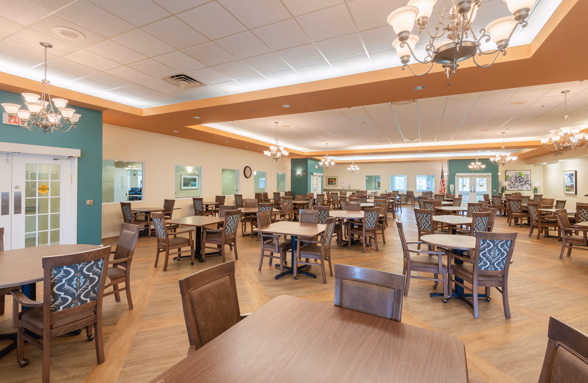 Spacious dining room with multiple wooden tables and chairs arranged neatly. The room features a warm color palette with teal and beige walls, chandeliers hanging from the ceiling, and large windows allowing natural light to enter. The floor has a wood pattern, and there are framed pictures on the walls.