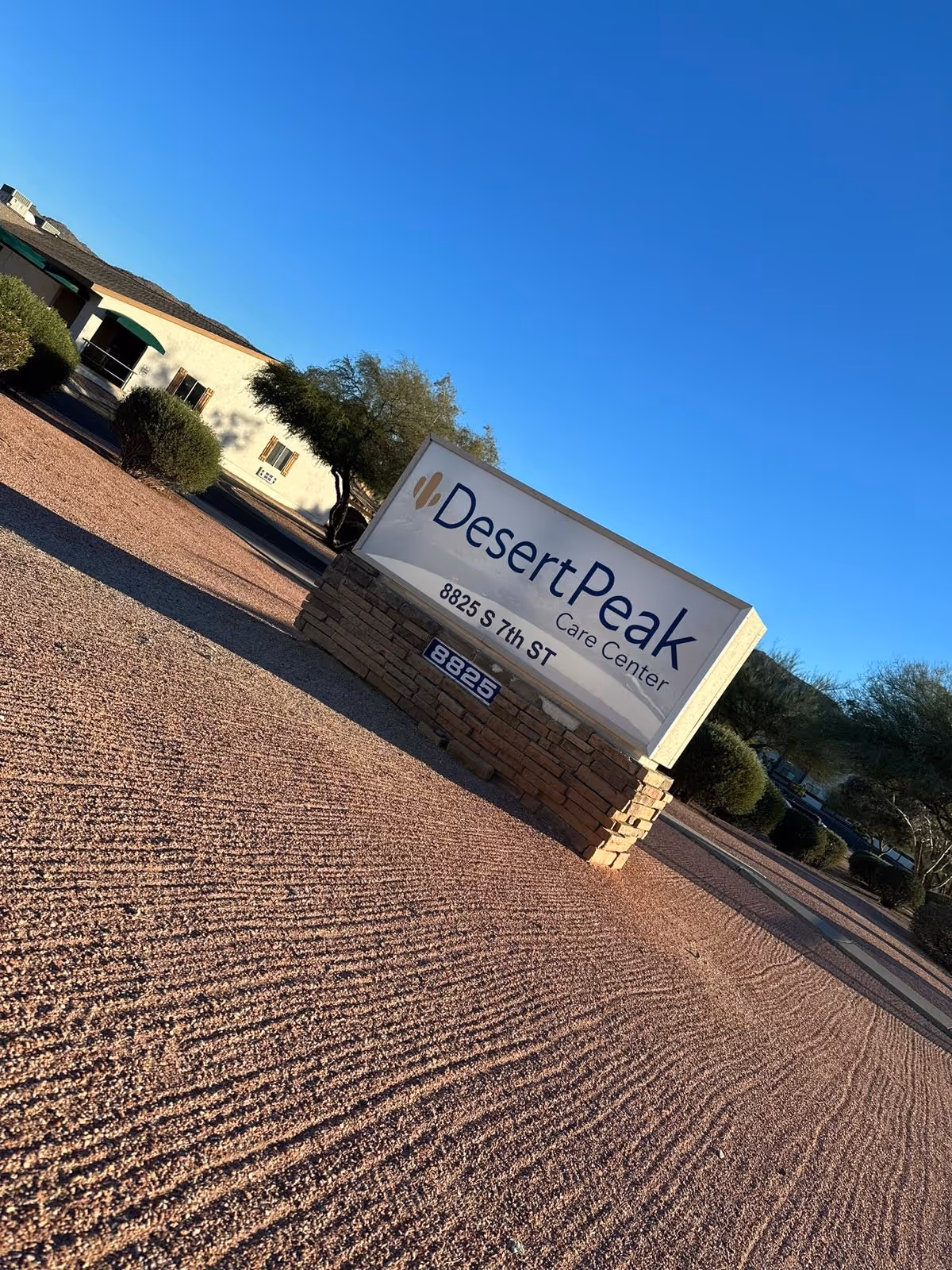 Outdoor view of the Desert Peak Care Center sign with the address 8825 S 7th St, surrounded by desert landscaping with gravel and bushes, under a clear blue sky.