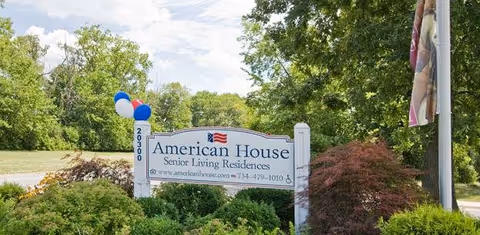 Entrance sign reading "American House Senior Living Residences" surrounded by bushes and trees with balloons attached.