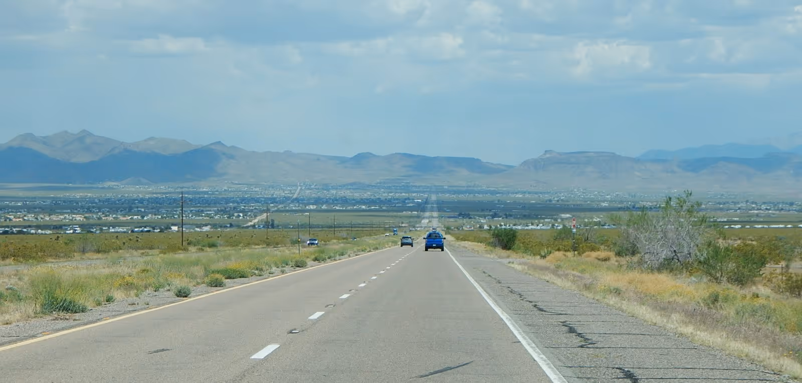 A desert highway stretches to distant mountains under a blue cloudy sky.
