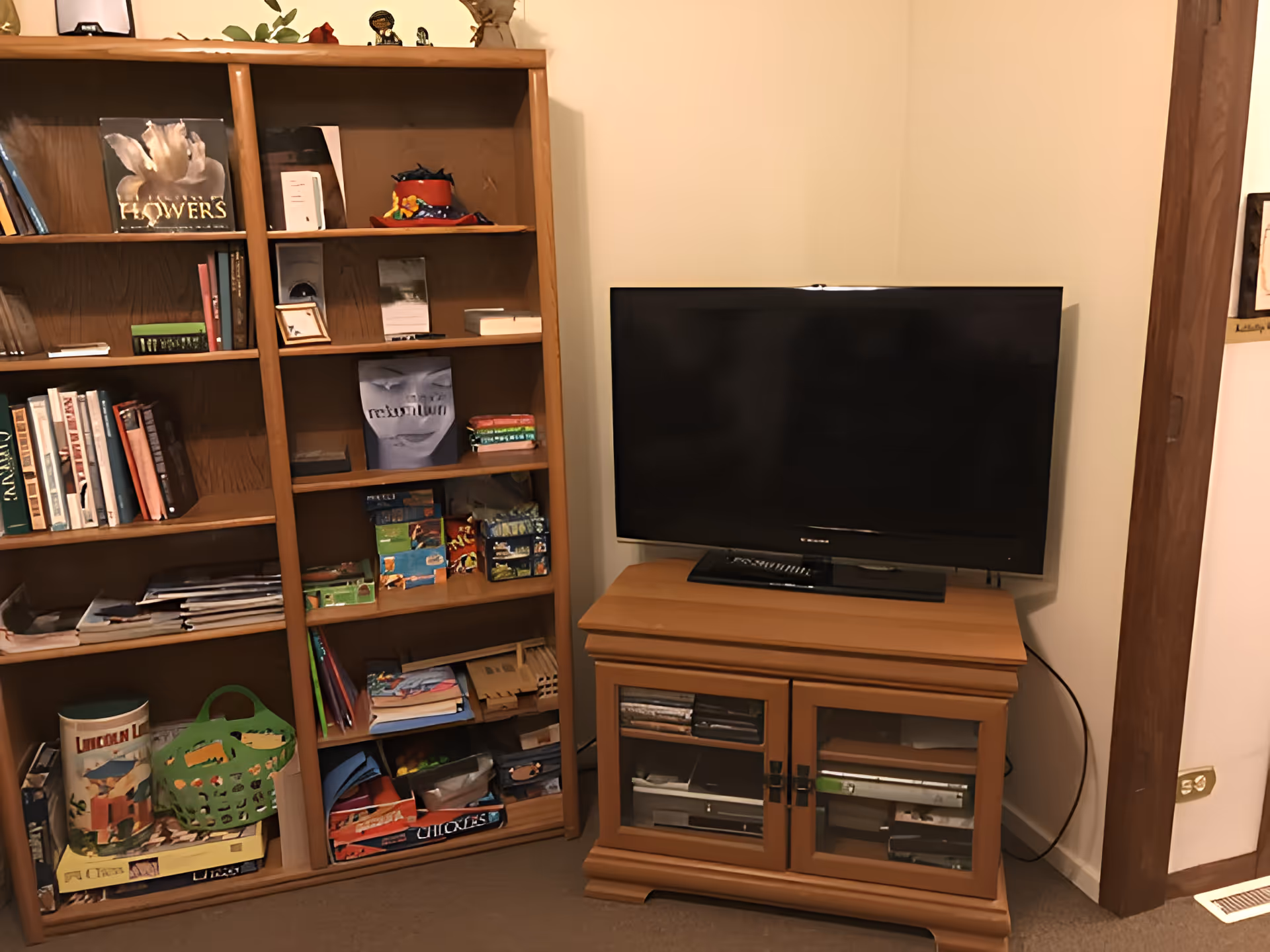 A cozy corner of a room featuring a wooden bookshelf filled with books, games, and decorative items next to a wooden TV stand with a flat-screen television on top. The TV stand has glass doors revealing electronic devices inside. The walls are light-colored, and there is a power outlet and vent visible near the floor.