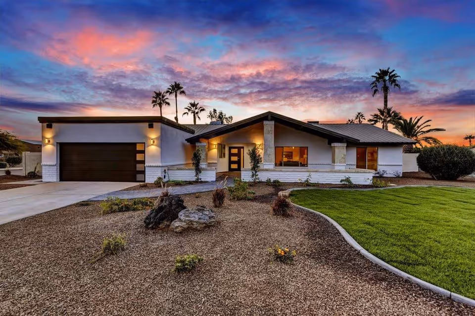Exterior view of a modern single-story house at sunset with a well-maintained lawn, desert landscaping with rocks and small plants, and palm trees in the background under a colorful sky.