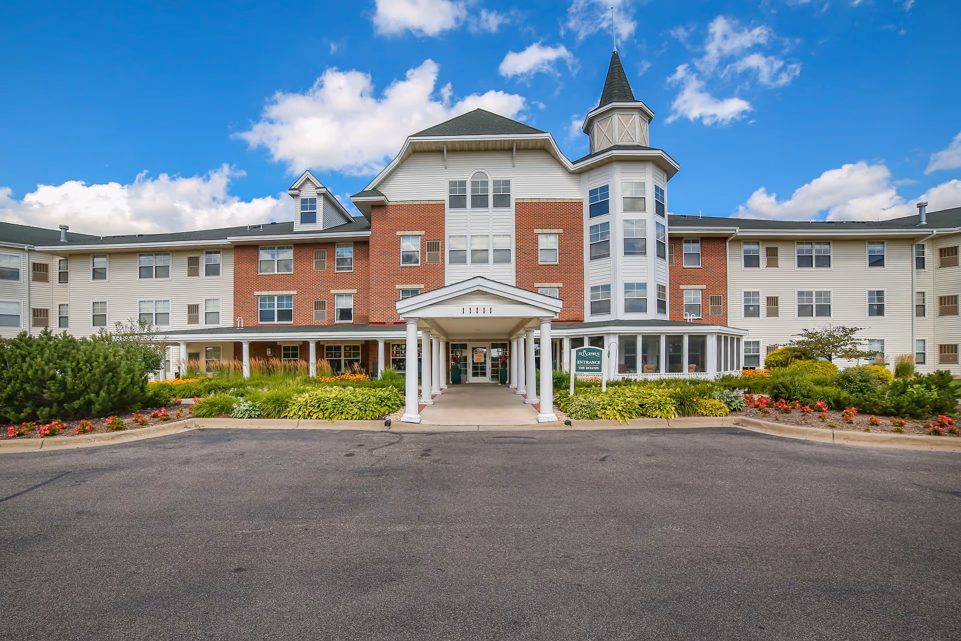 Front exterior view of The Rivers Retirement Community building with a covered entrance, well-maintained landscaping including bushes and flowers, and a blue sky with some clouds in the background.