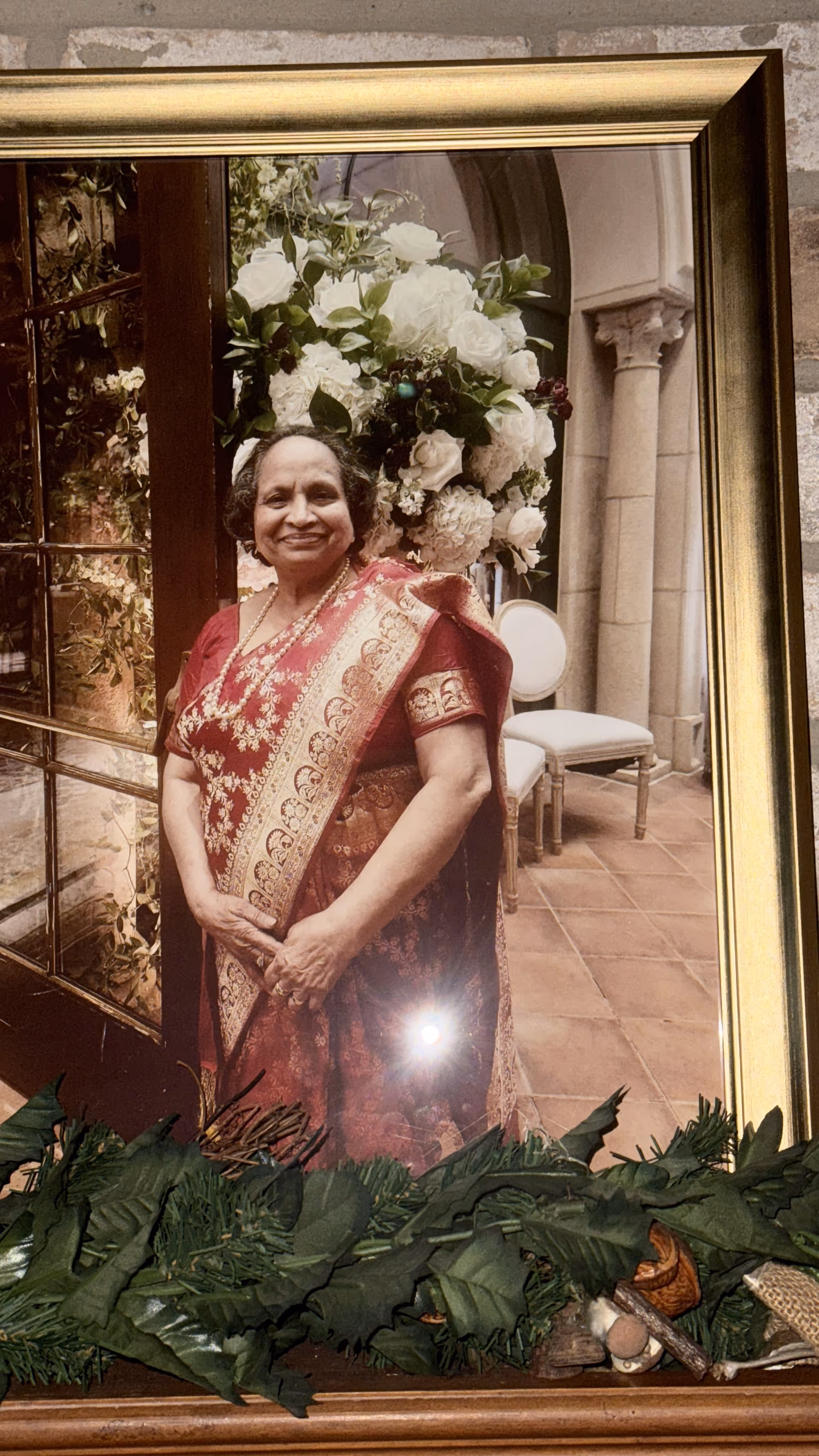 Framed portrait of a smiling woman in a red sari with white flowers behind