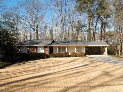Single-story brick house with a large front yard and driveway, surrounded by tall trees without leaves under a clear blue sky.