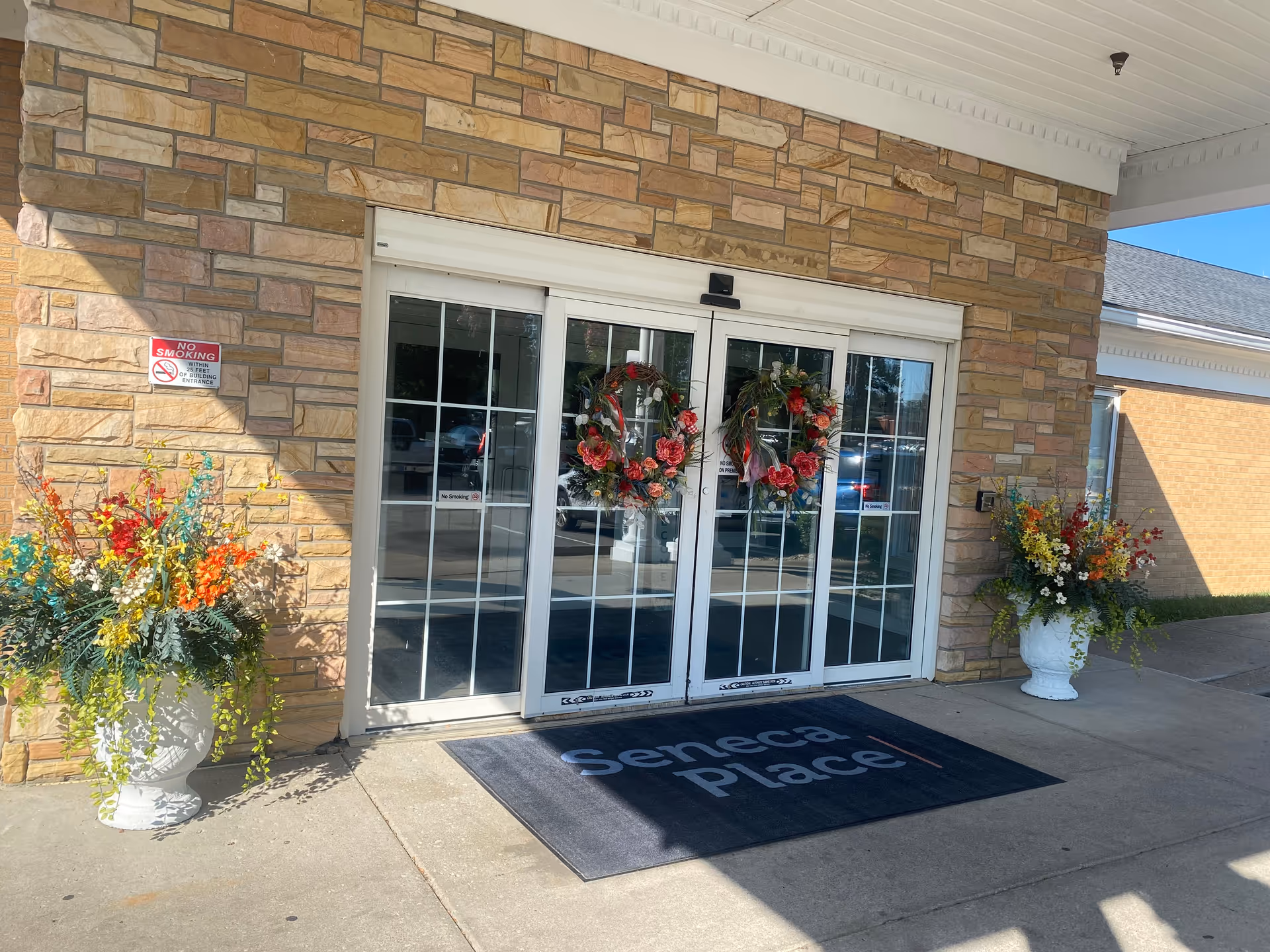 Entrance to Seneca Place featuring glass doors, wreaths, and planters against a stone facade.