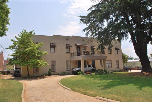 Exterior view of a two-story beige building with multiple windows and a balcony. There is a driveway curving in front of the building, a white vehicle parked under the balcony, and large trees and green grass surrounding the area.