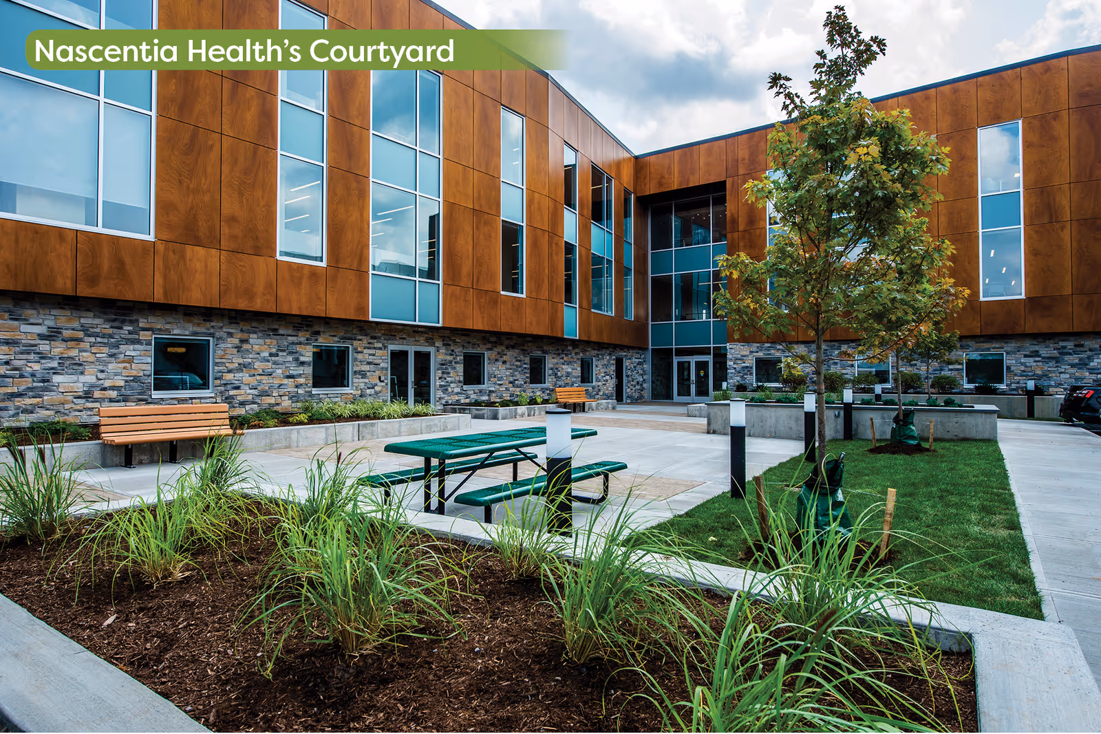 Nascentia Health courtyard featuring benches, picnic tables, trees, and modern brick-and-metal building.