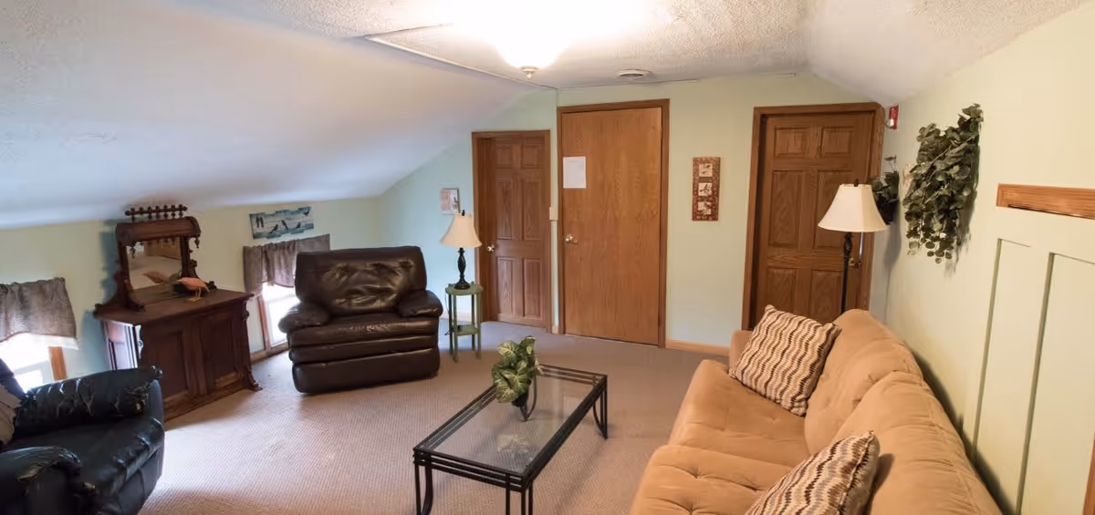 A cozy living room with a beige sofa adorned with patterned pillows, two dark leather armchairs, a glass coffee table, two wooden doors, a wooden dresser with a mirror, two table lamps, and wall decorations including framed art and greenery.