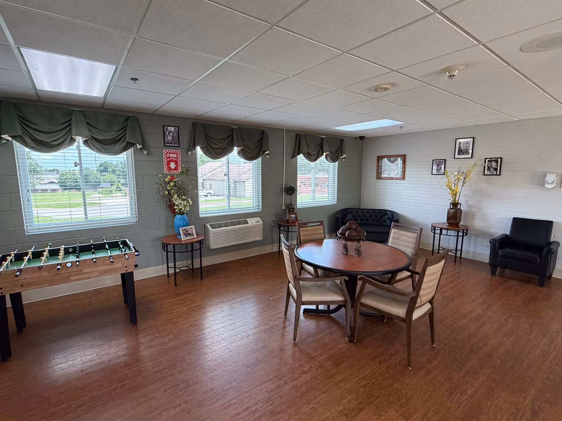Senior living facility lounge with round table, seating, foosball table, and windows.