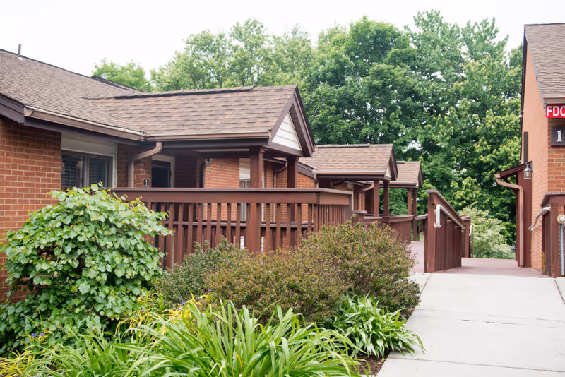 Exterior walkway and covered porches of a brick senior living building with wooden railings and landscaped greenery.