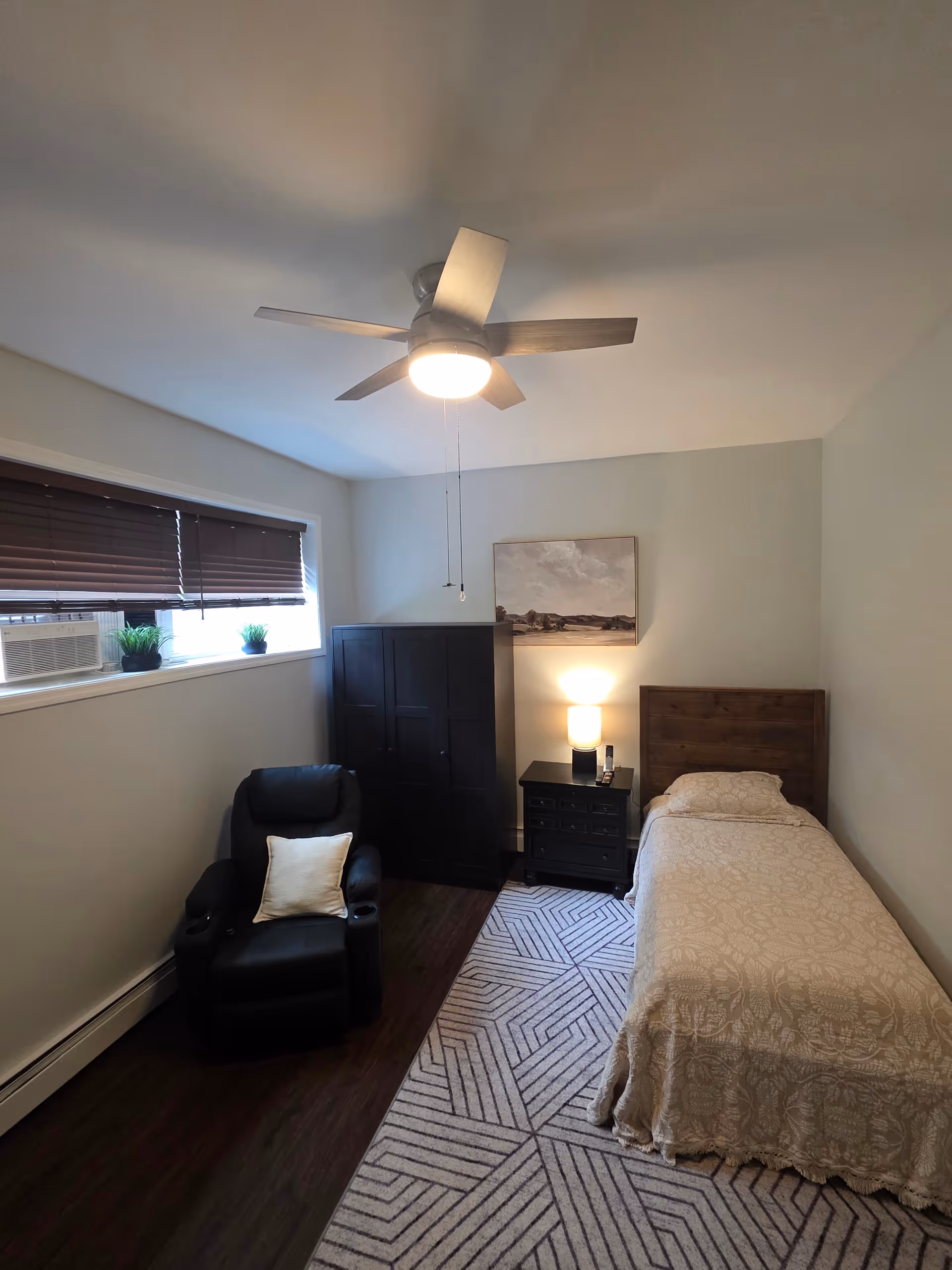 A small bedroom with a single bed, dresser, nightstand, recliner, window, and ceiling fan.