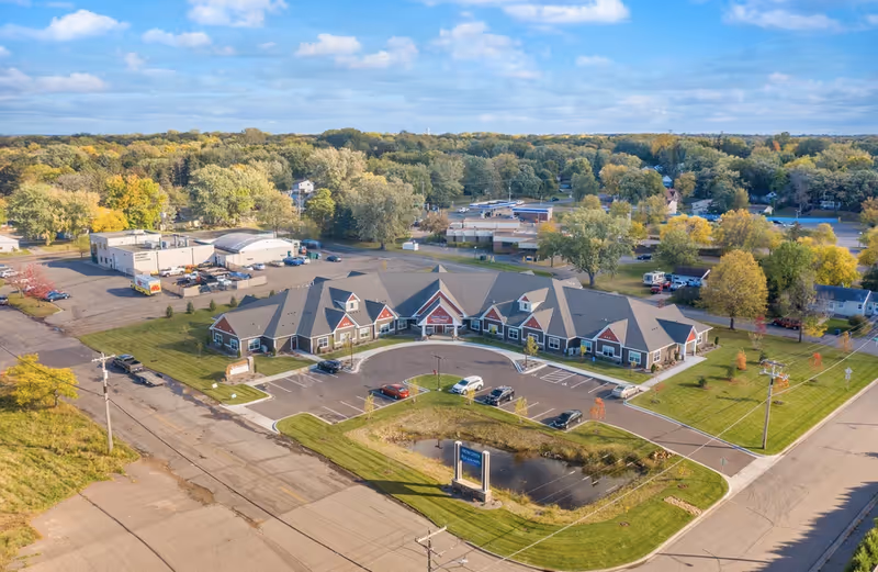 Aerial view of a single-story senior living building with a semicircular front driveway, parking lot, small pond and surrounding trees.