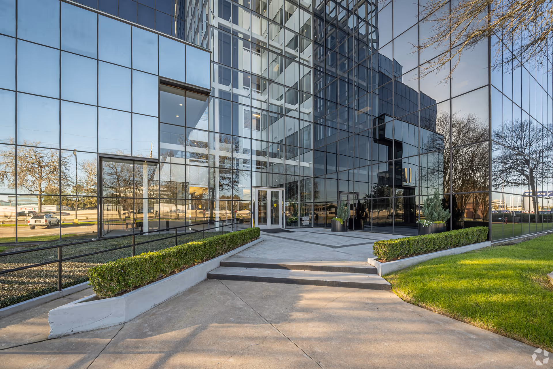 Glass-fronted building entrance with steps, trimmed hedges, and reflective windows