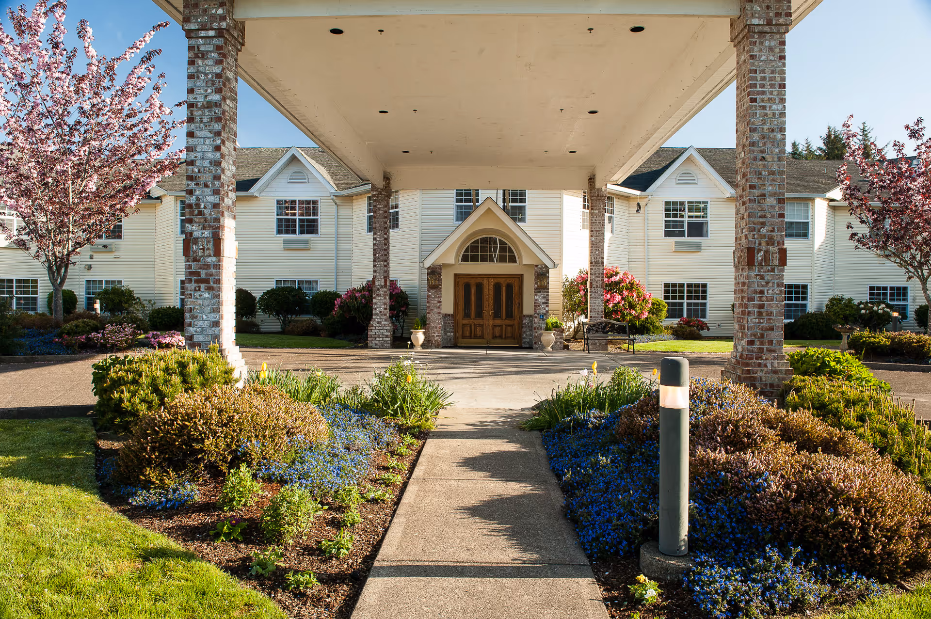 Front entrance of a senior living building with a covered porte-cochère, walkway and landscaped flower beds.
