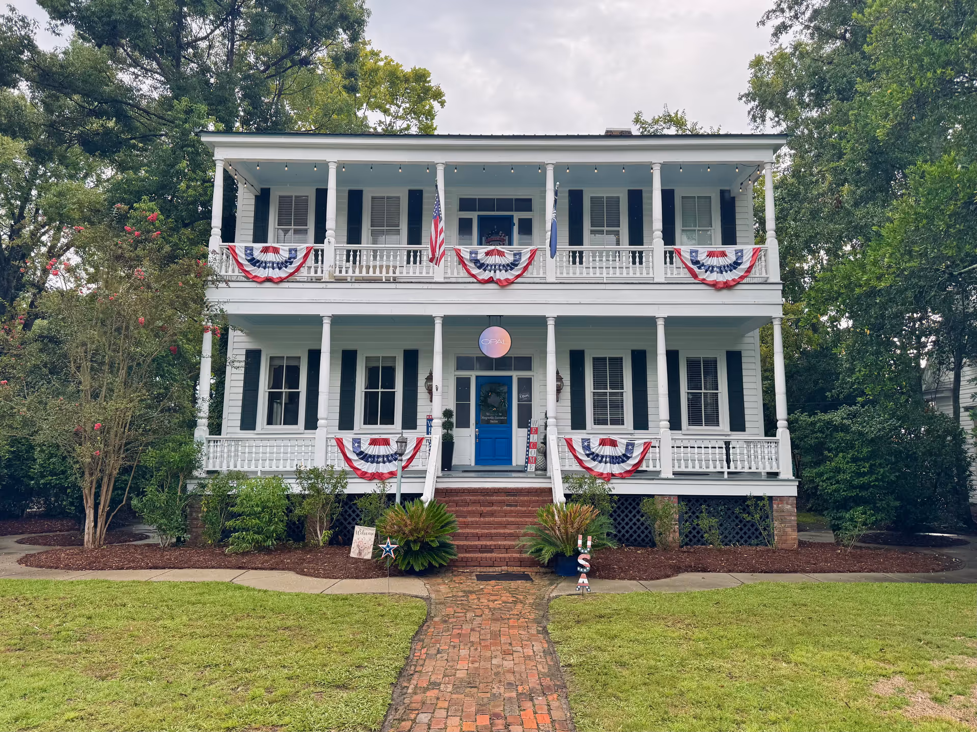 Two-story white front-porch building with blue shutters, brick steps, and patriotic bunting.