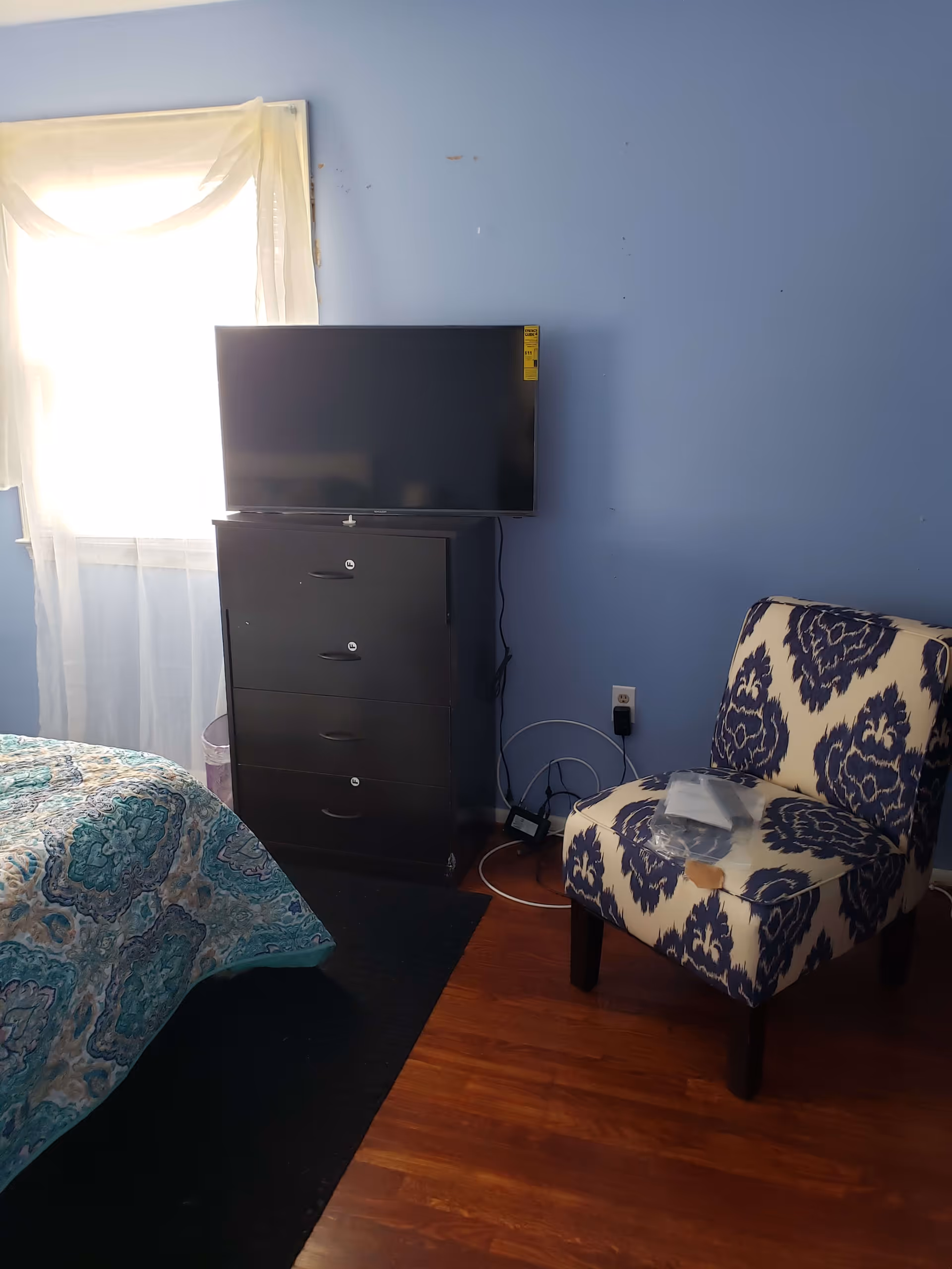 A bedroom with blue walls and wooden floor. There is a bed with a blue and green patterned quilt partially visible on the left. Next to the bed is a black dresser with four drawers, on top of which sits a flat-screen TV. To the right of the dresser is a patterned upholstered chair with blue and white designs, and some plastic-wrapped items placed on the seat. A window with sheer white curtains is behind the dresser, letting in bright natural light.