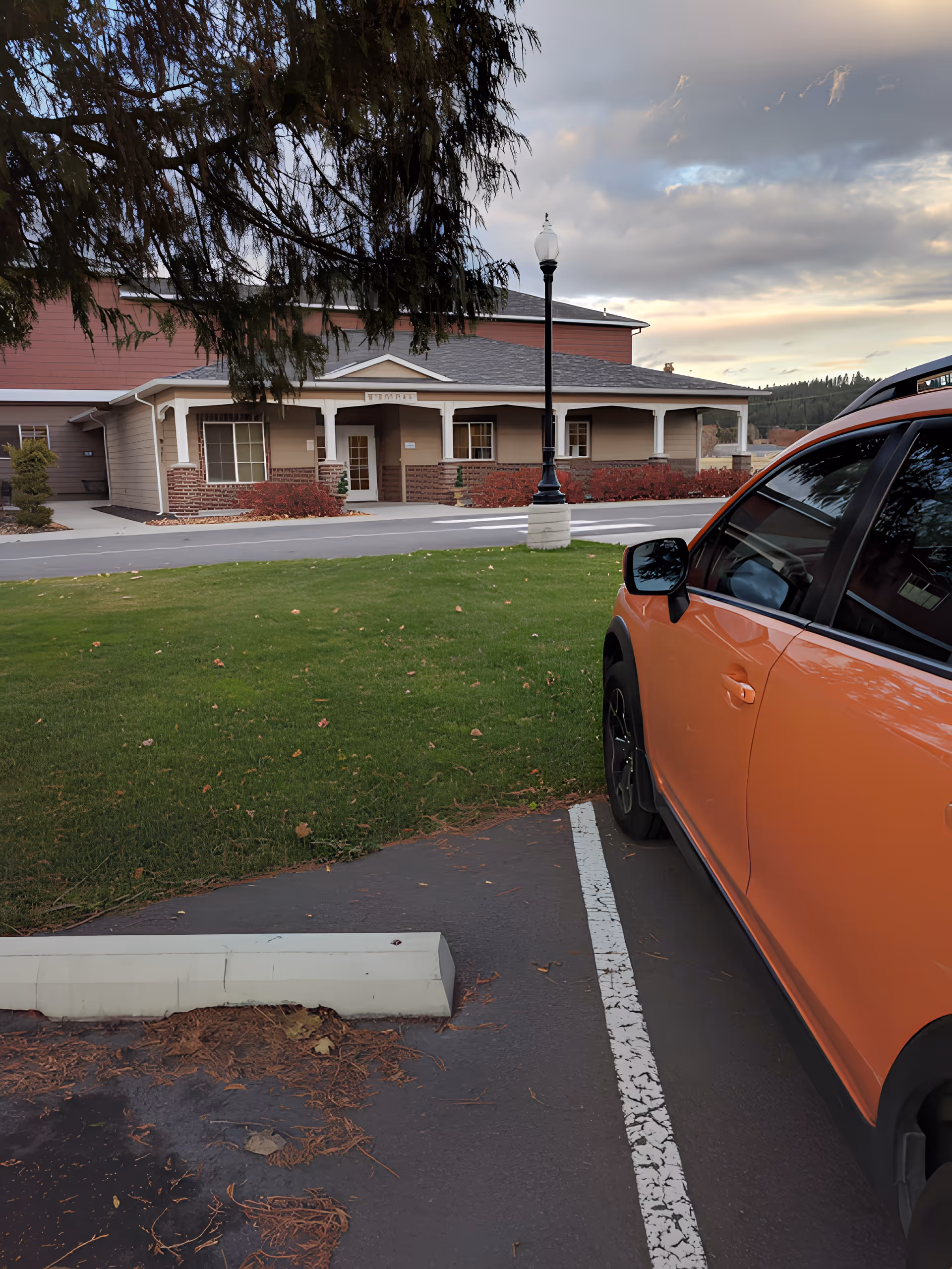 View of the exterior of a building with a covered porch and brick accents, a green lawn in front, a street lamp, and an orange car parked in a parking space in the foreground under a cloudy sky.