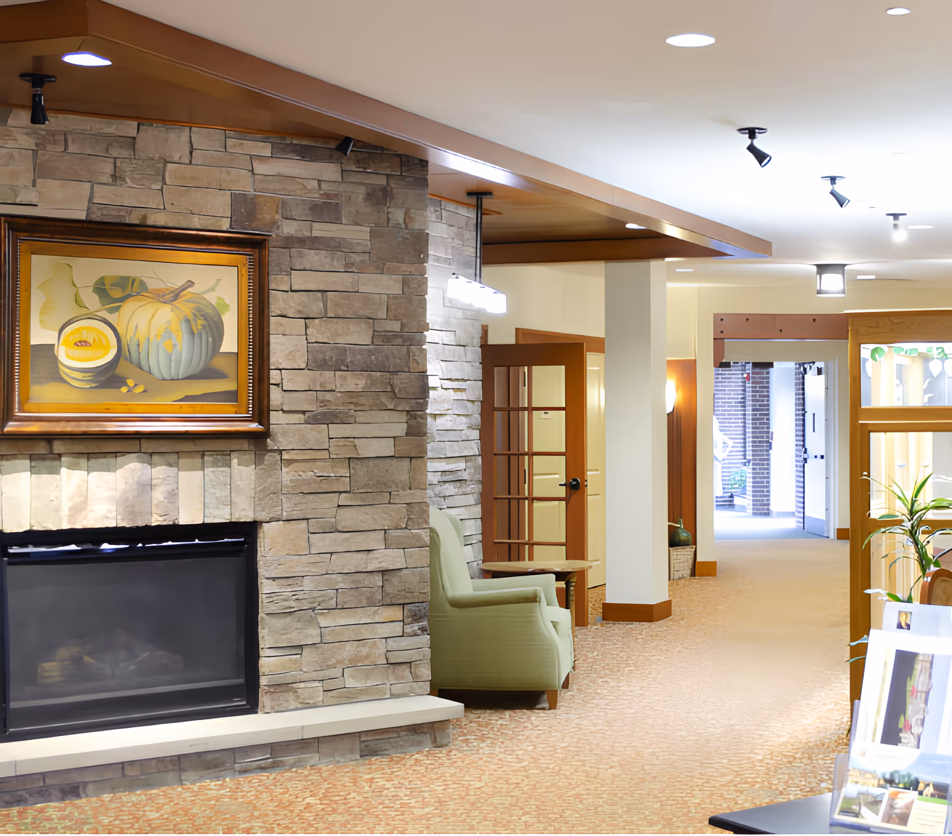 Interior common area of a senior living facility with a stone fireplace, framed artwork, seating and a hallway.