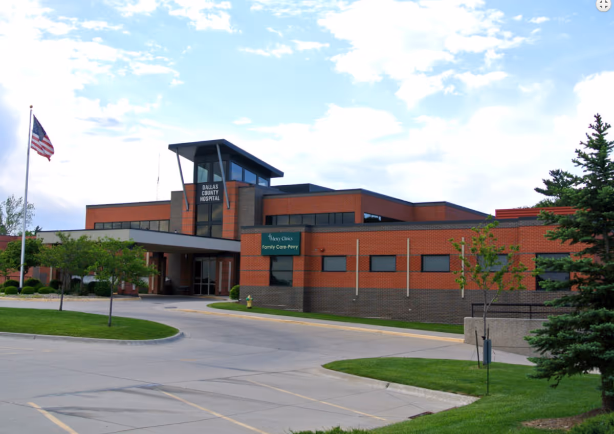 Exterior view of a modern brick building with a sign that reads Dallas County Hospital and Mercy Clinics Family Care-Perry. The building is surrounded by a paved driveway, green grass, trees, and an American flag on a flagpole. The sky is partly cloudy.