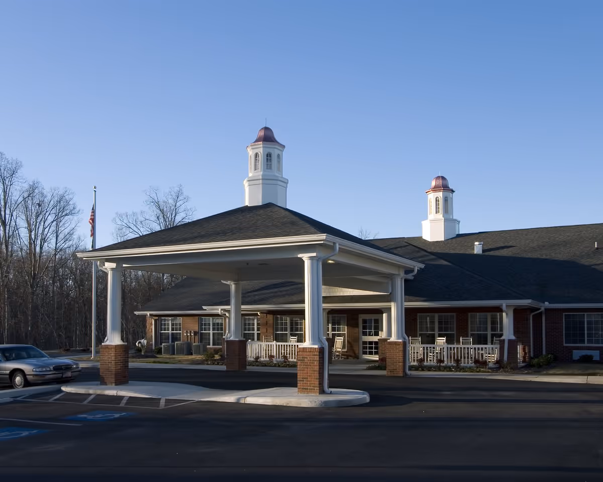 Exterior view of a single-story assisted living facility building with a covered entrance supported by white columns with brick bases. The building has two cupolas on the roof and several windows. There is a parked car and an American flag on a flagpole near the building. Trees without leaves are visible in the background under a clear blue sky.