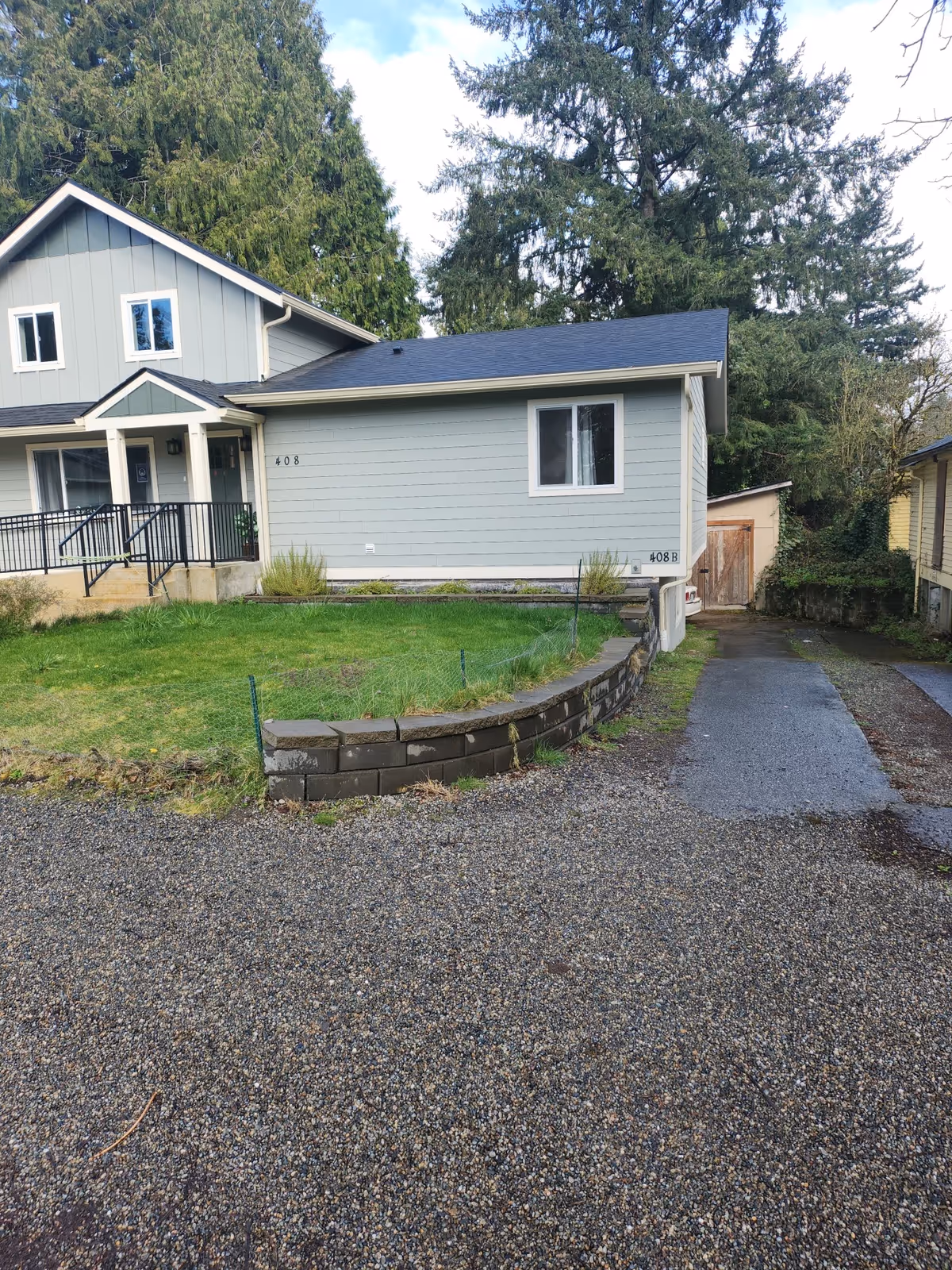 Exterior view of a light gray residential building with front porch and gravel driveway.
