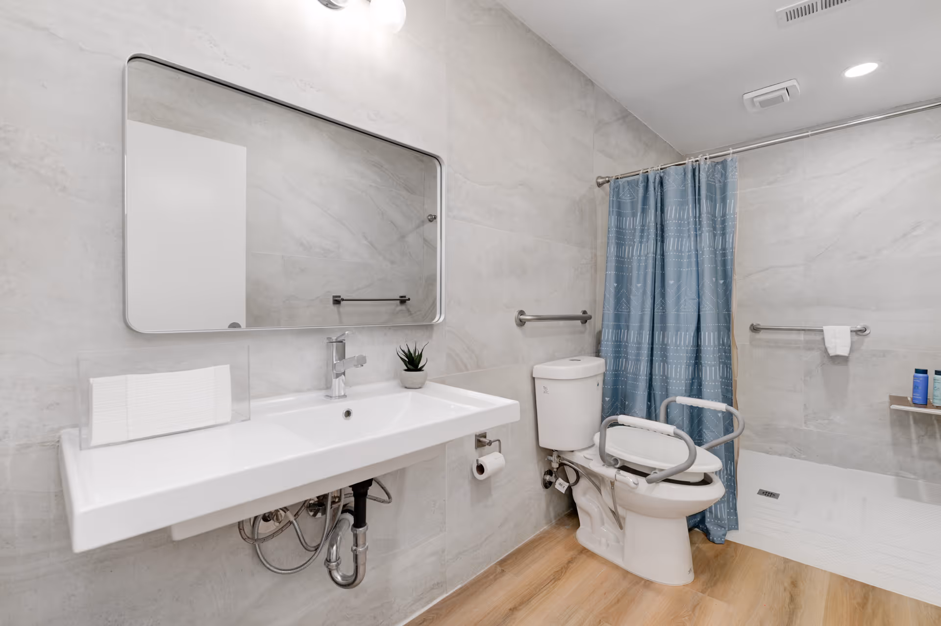 Accessible bathroom with sink, mirror, toilet, grab bars, and blue shower curtain, tiled walls.