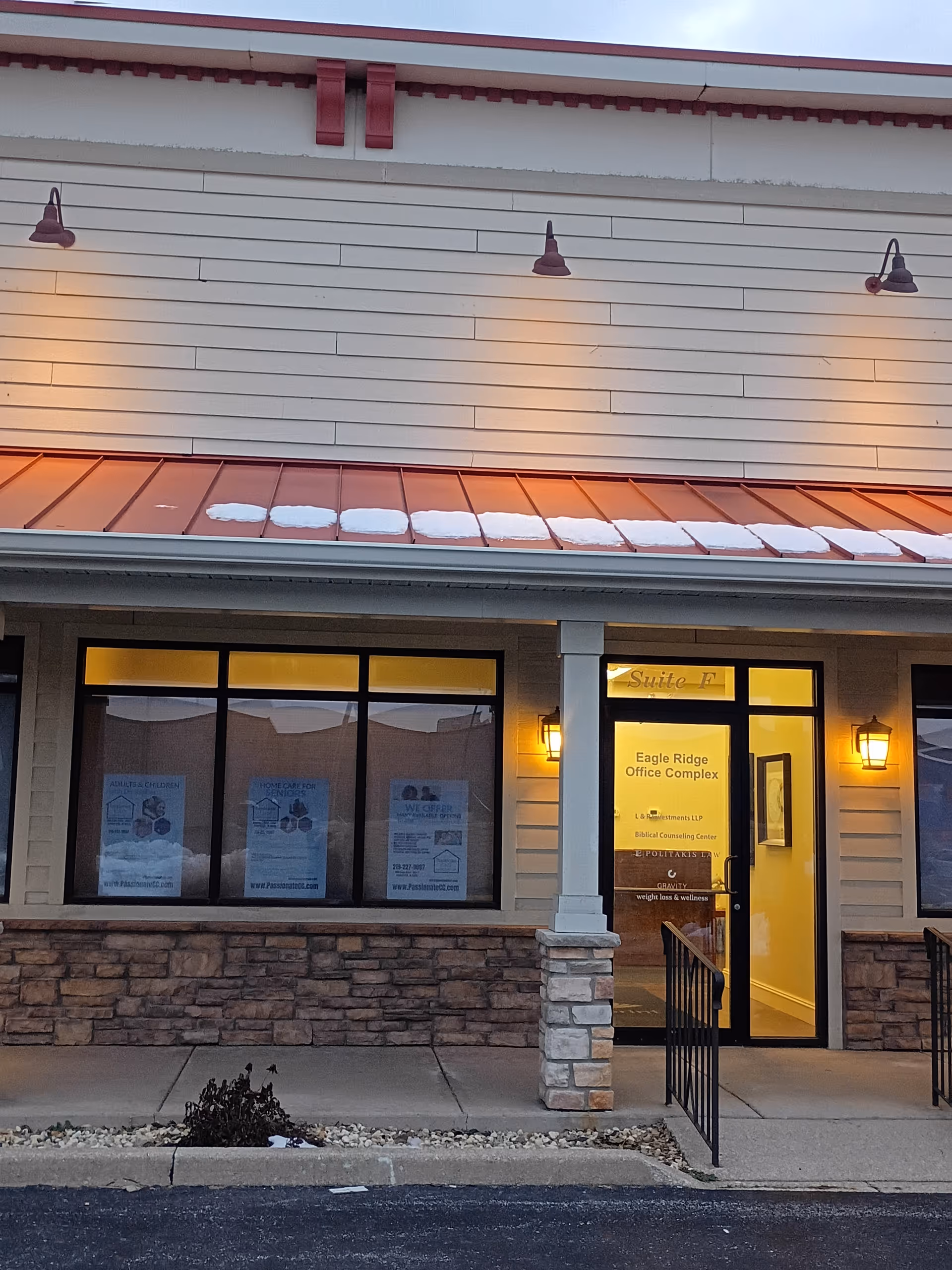 Exterior storefront with large windows, stone base, and illuminated entrance at dusk.