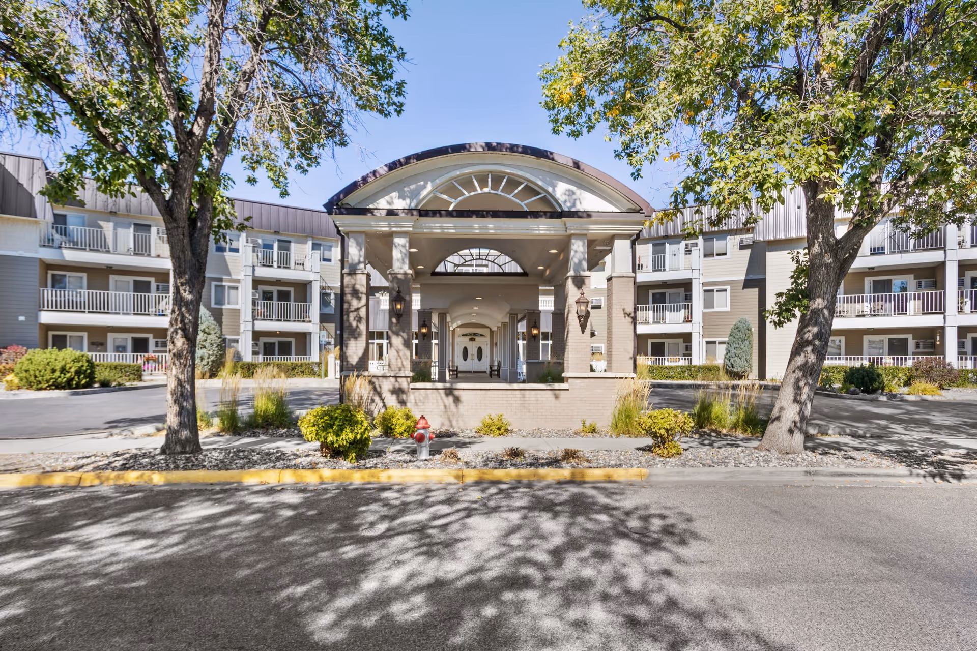 Front exterior view of Westpark Village senior living facility showing a covered entrance with columns, surrounded by trees and landscaping, and a multi-story building with balconies in the background under a clear blue sky.