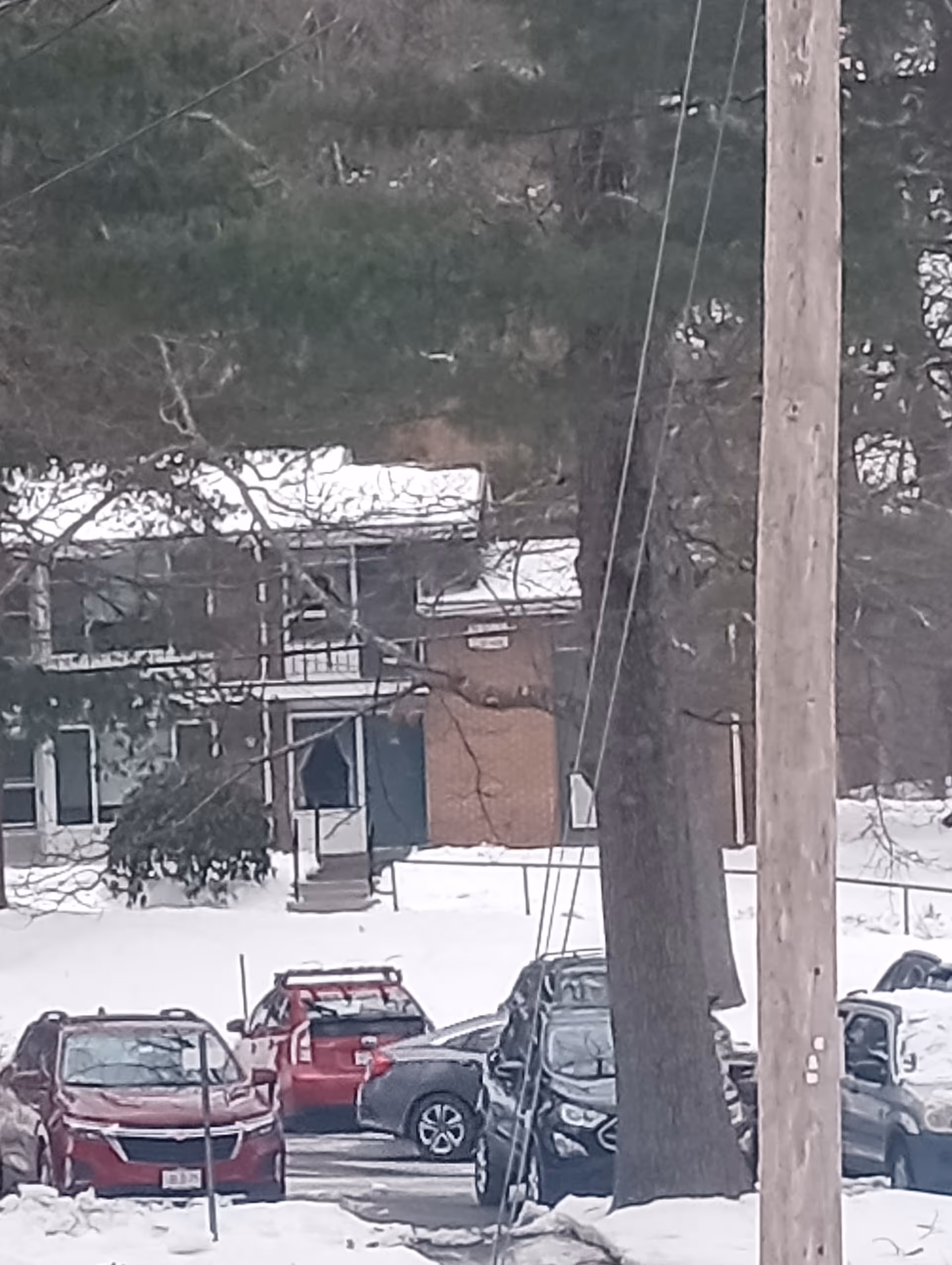 Snow-covered parking area in front of an elder care facility, with cars, trees, and utility pole.