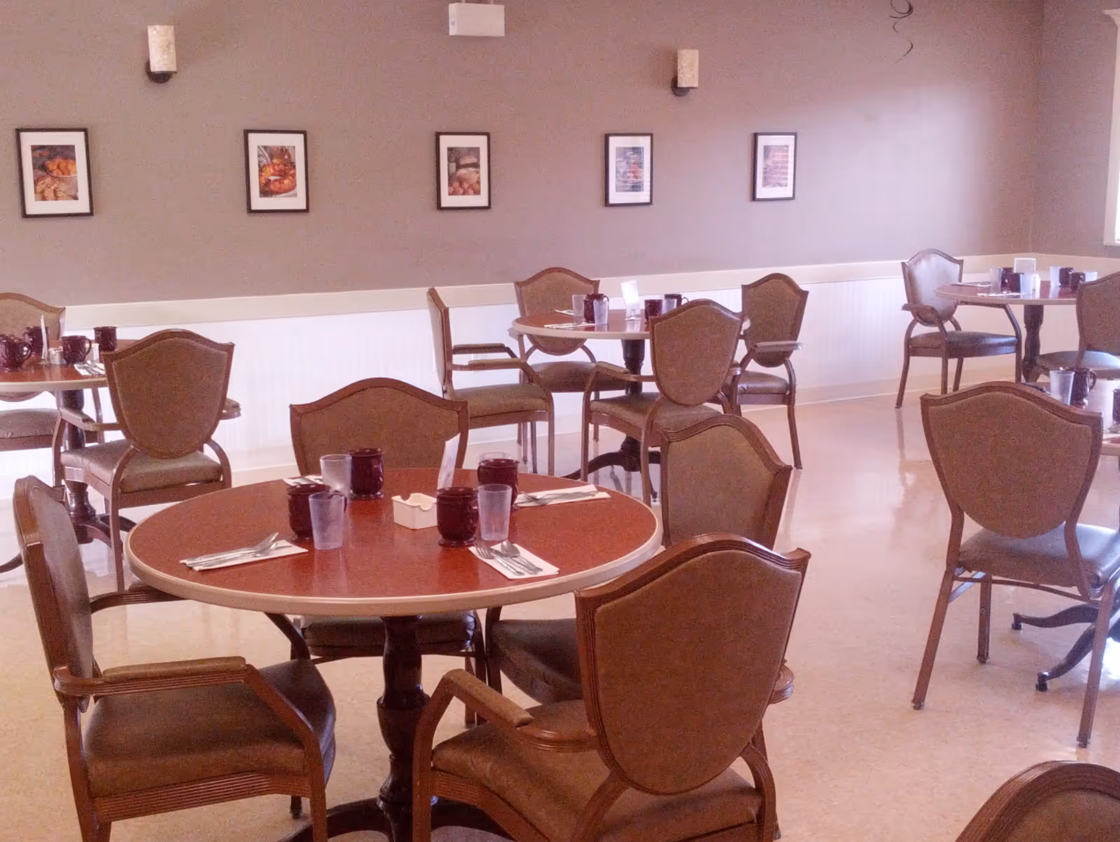 A dining room with several round wooden tables and cushioned chairs arranged around them. Each table is set with purple drinking glasses, silverware wrapped in napkins, and small condiment holders. The walls are painted a muted brown with white wainscoting, decorated with framed pictures of food. The room is well-lit with natural light coming from a window on the right.