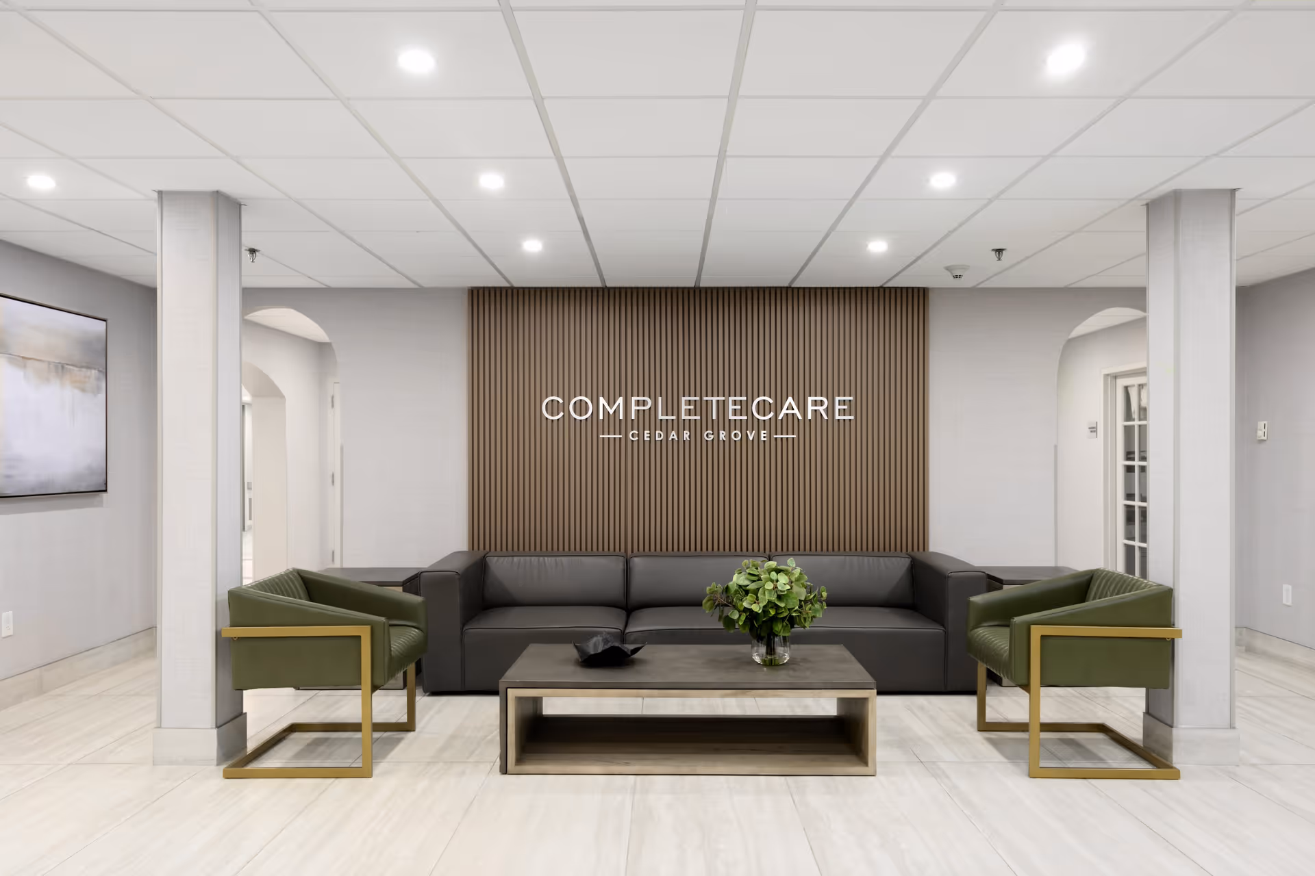 A modern and clean lounge area with a dark gray sectional sofa, two green armchairs with gold frames, and a rectangular coffee table with a plant centerpiece. The back wall features vertical wooden slats and a sign that reads 'Complete Care Cedar Grove'. The room has light-colored tiled floors and a white ceiling with recessed lighting.