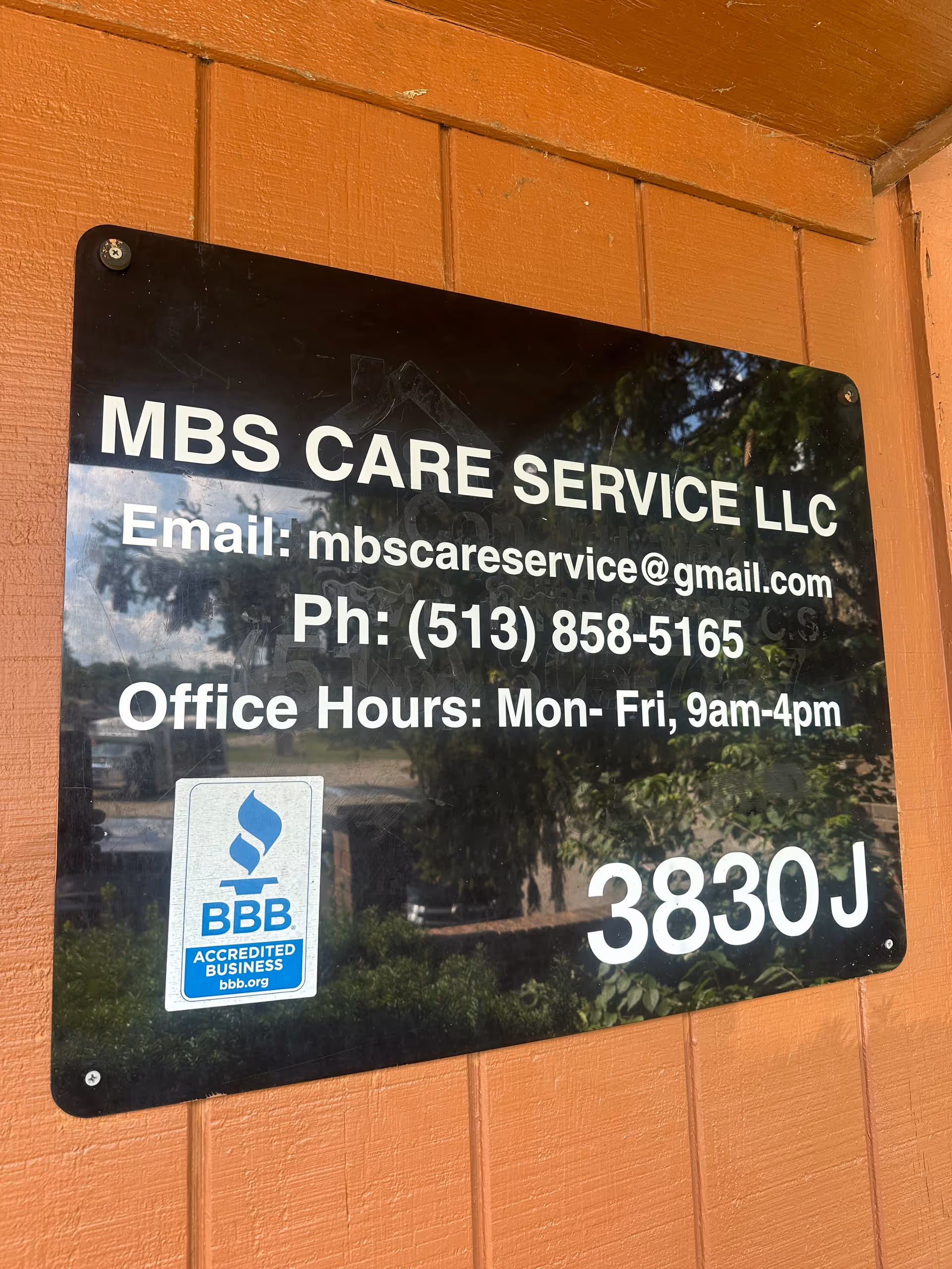 Black sign on orange wall displays MBS CARE SERVICE LLC contact information and hours.