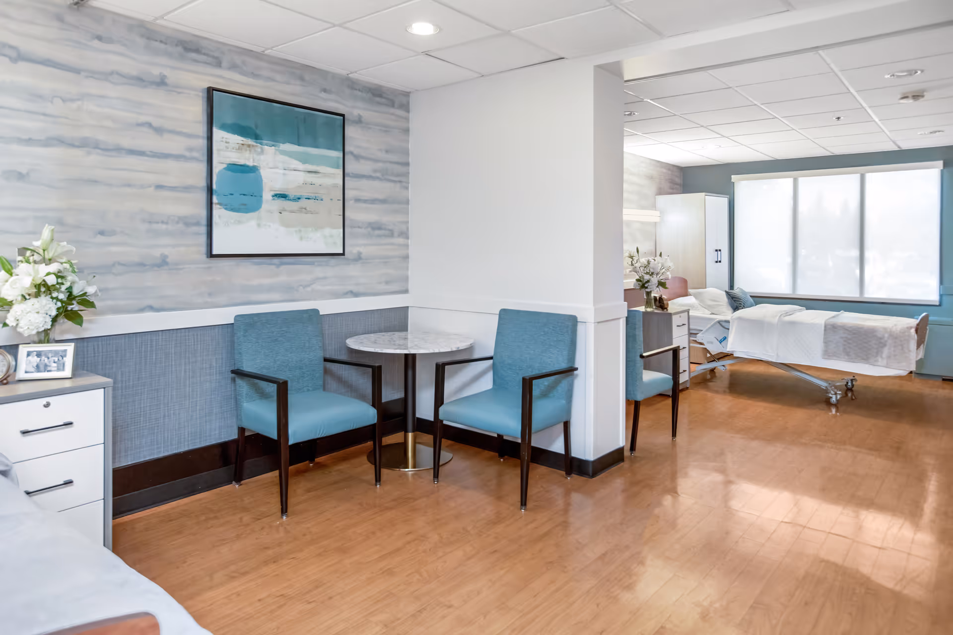Bright senior living lounge with blue chairs, round table, and a hospital bed in background.