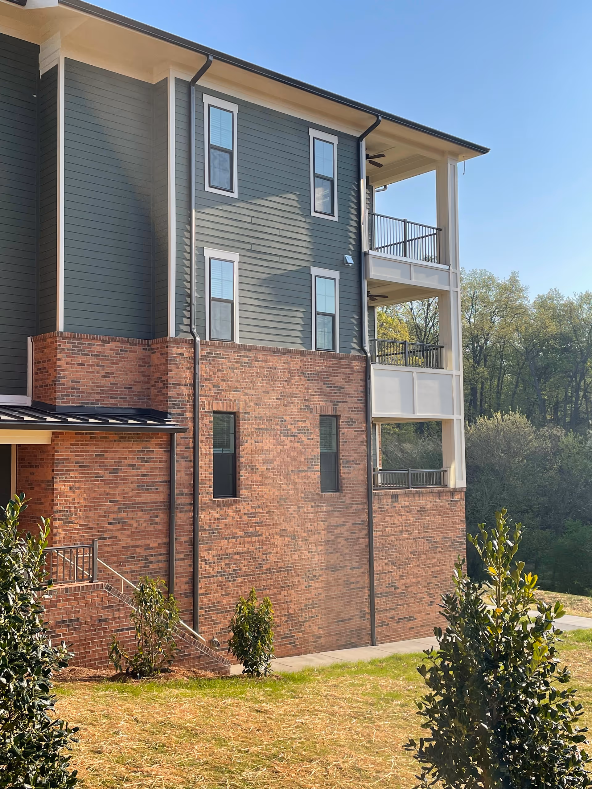 Exterior view of a multi-story building with a combination of red brick and green siding. The building has several windows and two covered balconies with railings. There are small bushes and a grassy area in the foreground, and trees in the background under a clear blue sky.
