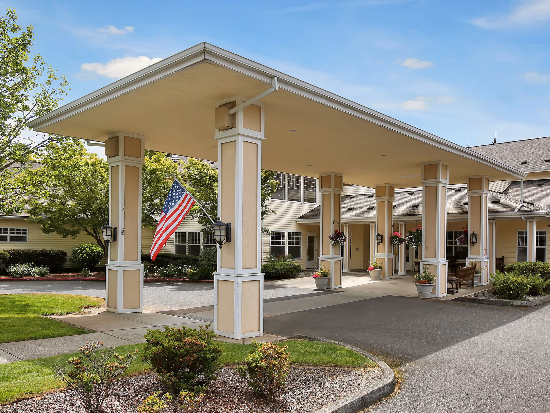 Entrance area of a senior living facility with a covered drop-off area supported by beige and white columns. There are hanging flower pots and potted plants around the entrance. An American flag is displayed on one of the columns. The building is light yellow with multiple windows and a well-maintained garden surrounding the driveway.