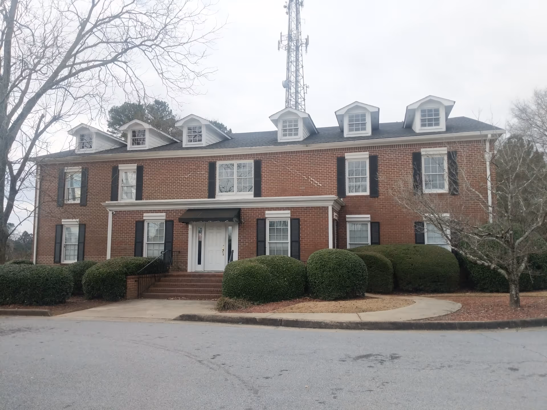 Brick senior living building with black shutters, dormer windows, and manicured hedges at entrance.