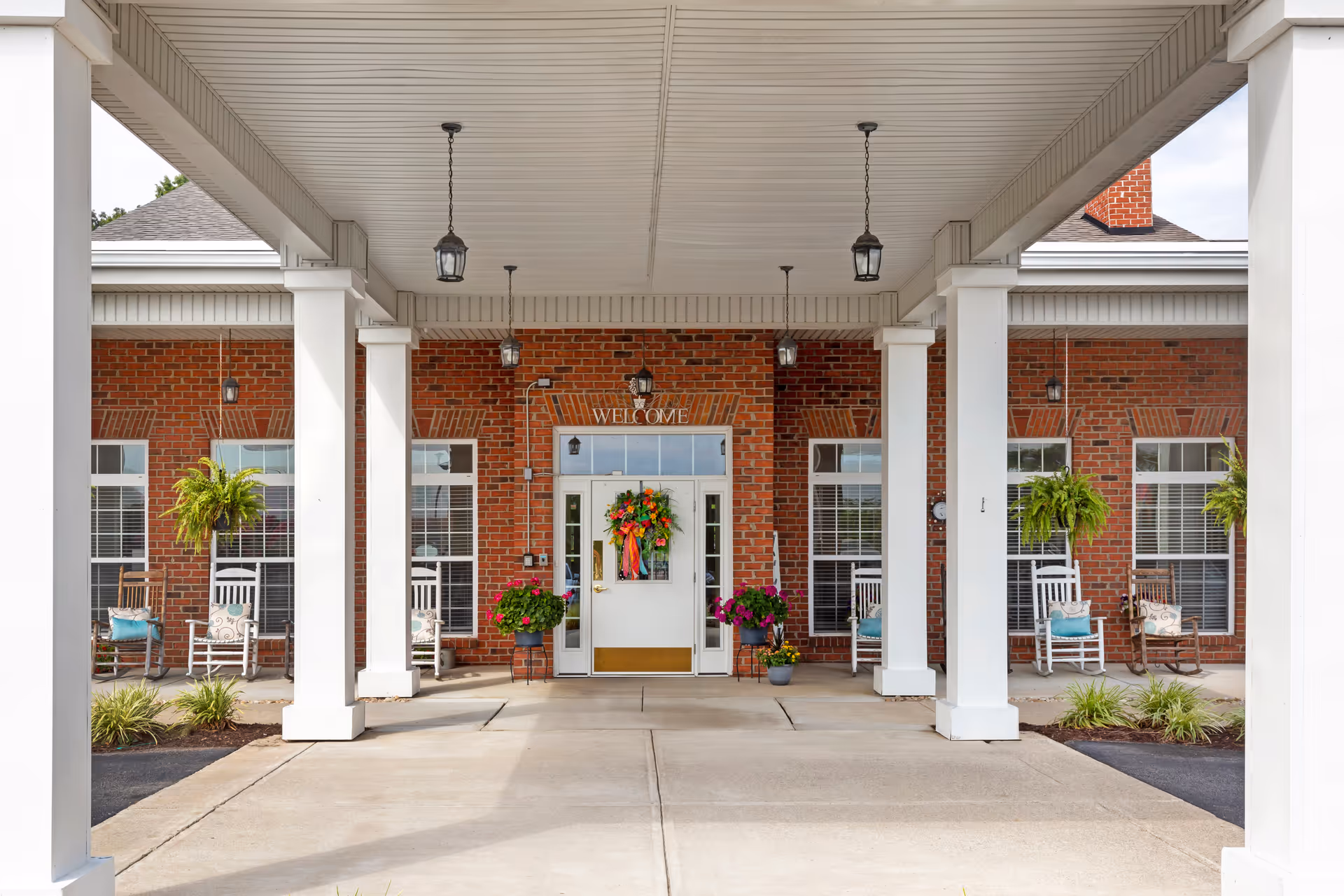 Brick building entrance with white columns, hanging lanterns, and potted plants on a covered walkway.