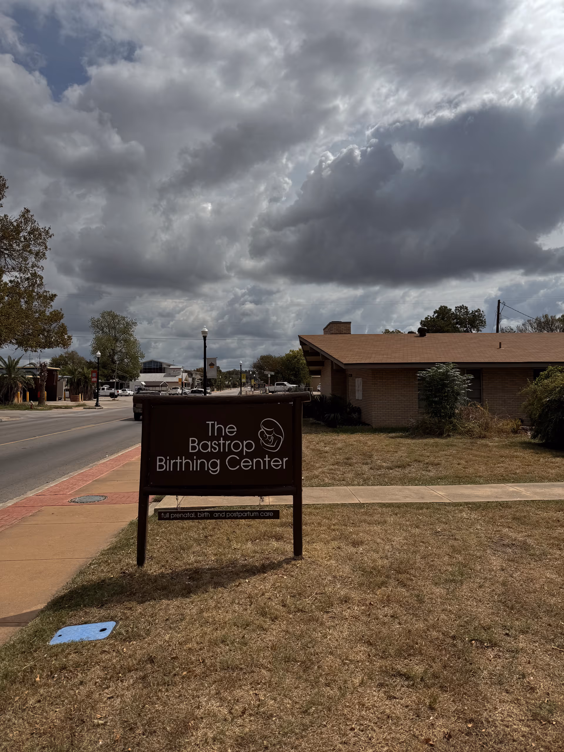 A wooden sign for The Bastrop Birthing Center beside a brick building along a street.