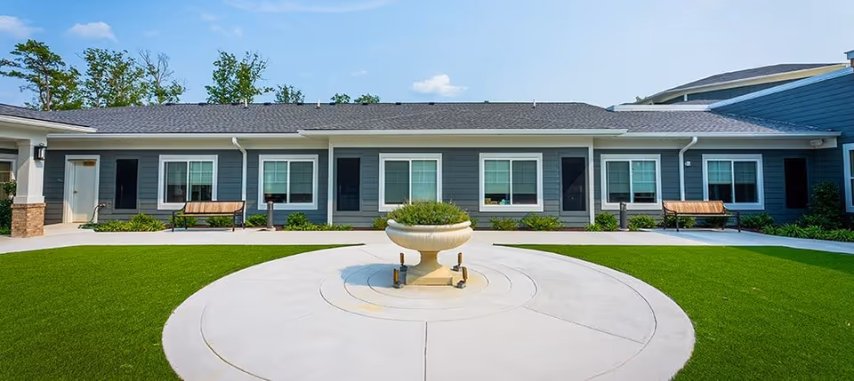 Front exterior of a single-story building with a circular walkway and central fountain surrounded by green lawn and benches.
