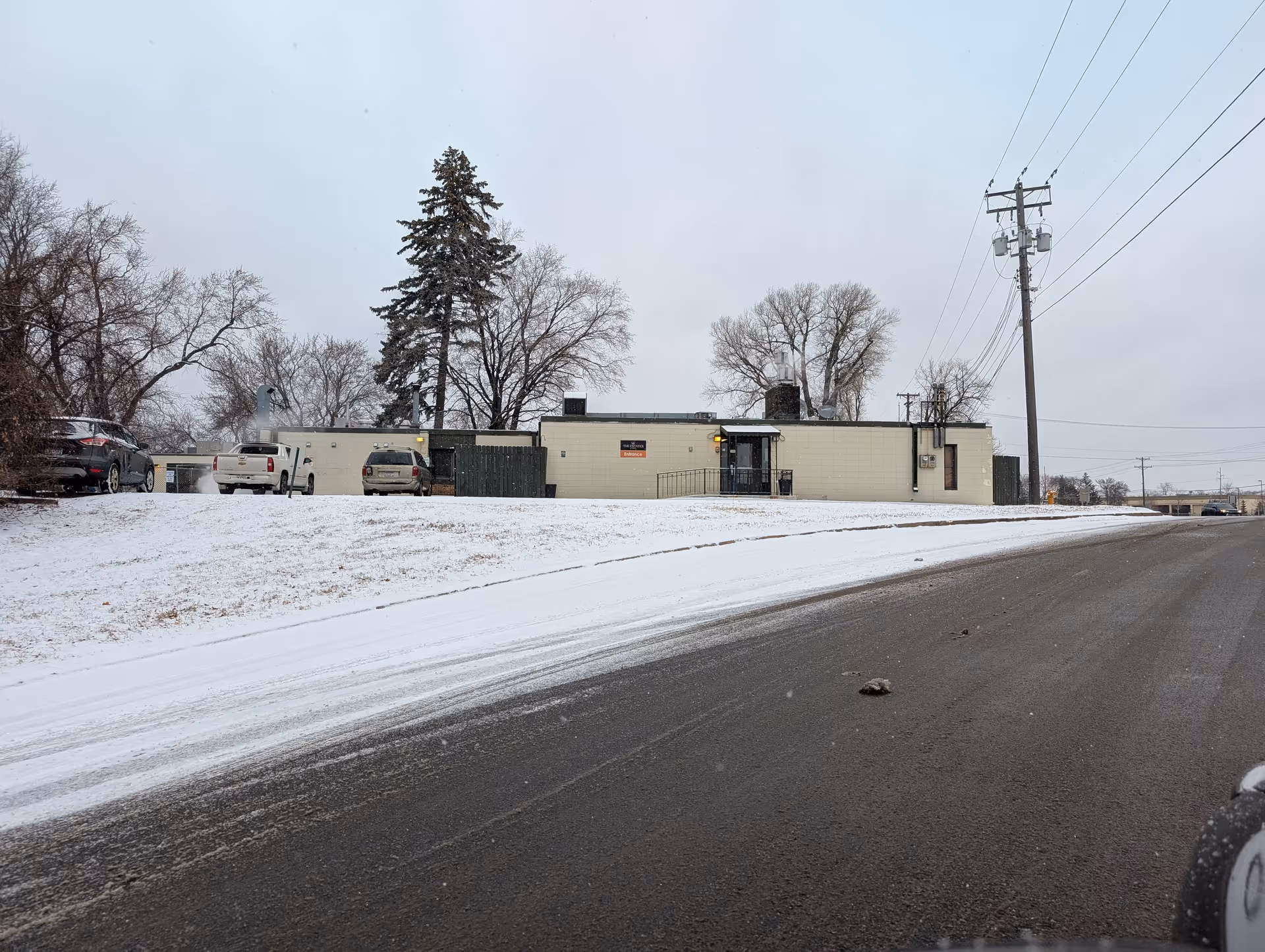 Single-story building with parked cars on a snow-covered lawn and power lines under a gray sky.
