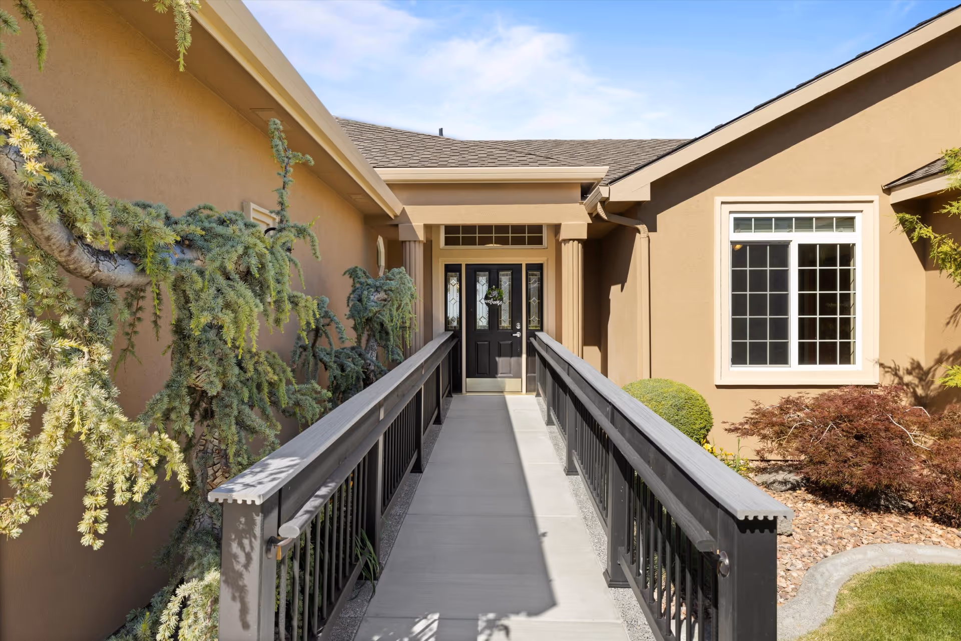 Entrance to Clovernook Adult Family Home with a concrete ramp and black railings leading to double doors, beige building and landscaped surroundings.