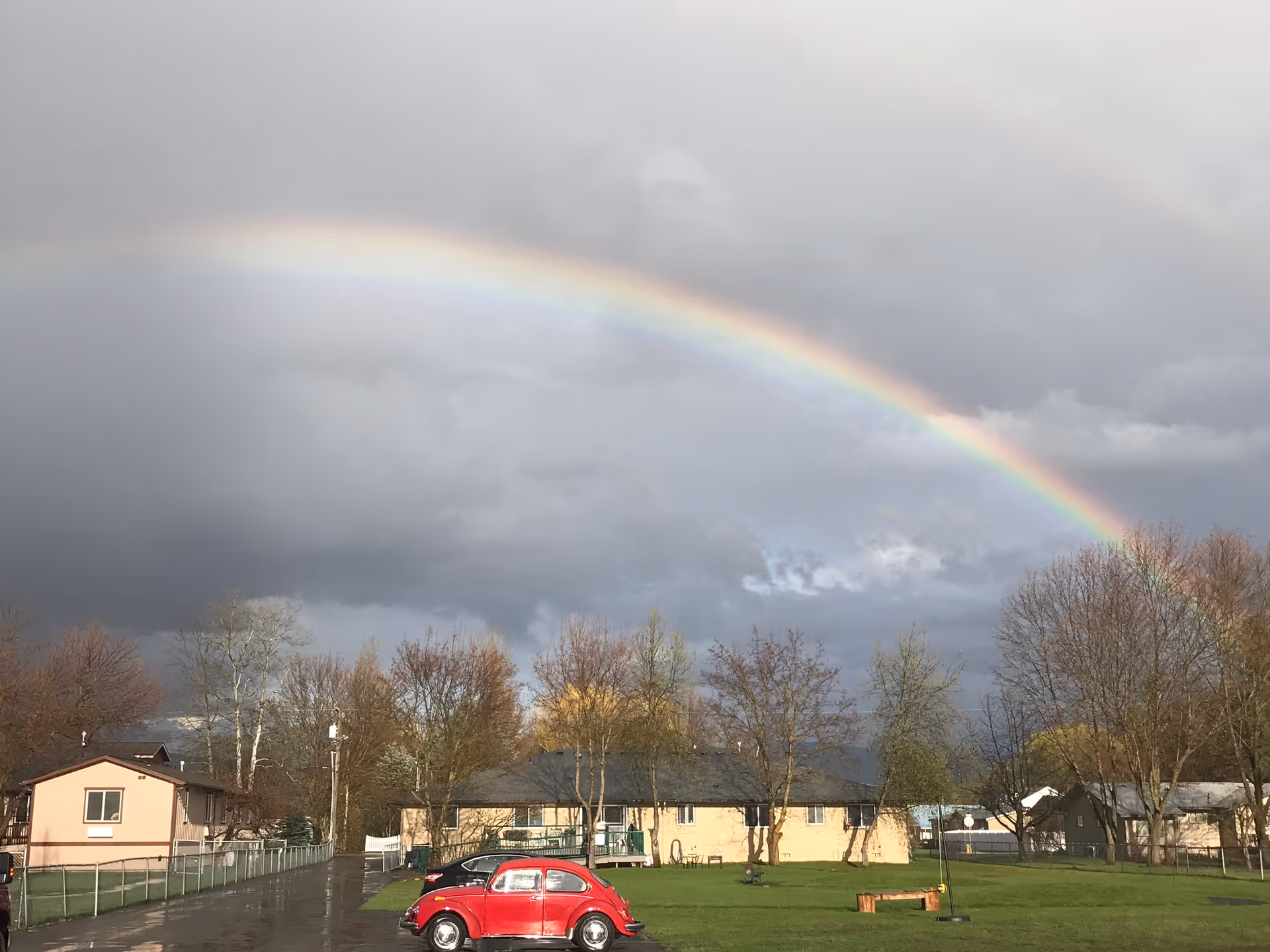 Rainbow arches over a grassy outdoor area with single-story homes and a red car.