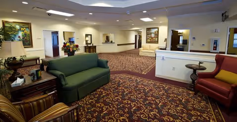 A spacious senior living facility common area with patterned carpet, multiple seating options including a green sofa, red armchair, and beige sofa. The room is decorated with framed paintings, a table lamp, and flower arrangements. The walls are light-colored, and there are several doorways leading to other rooms.