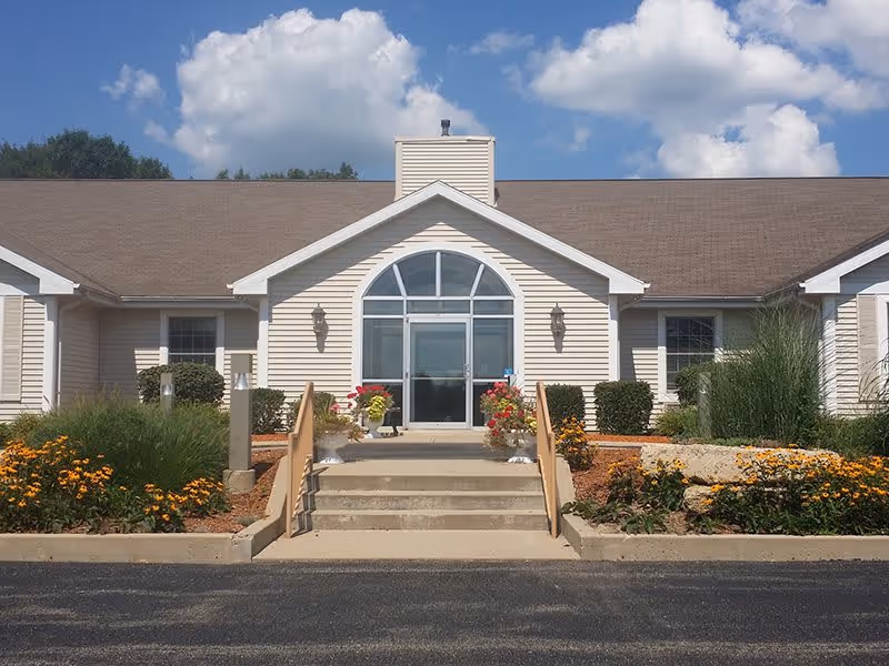 Front exterior view of a single-story building with beige siding and a brown roof under a blue sky with scattered clouds. There are steps leading up to a glass door entrance with an arched window above it, flanked by two wall-mounted lanterns. Flower beds with yellow and orange flowers and green shrubs line the walkway.